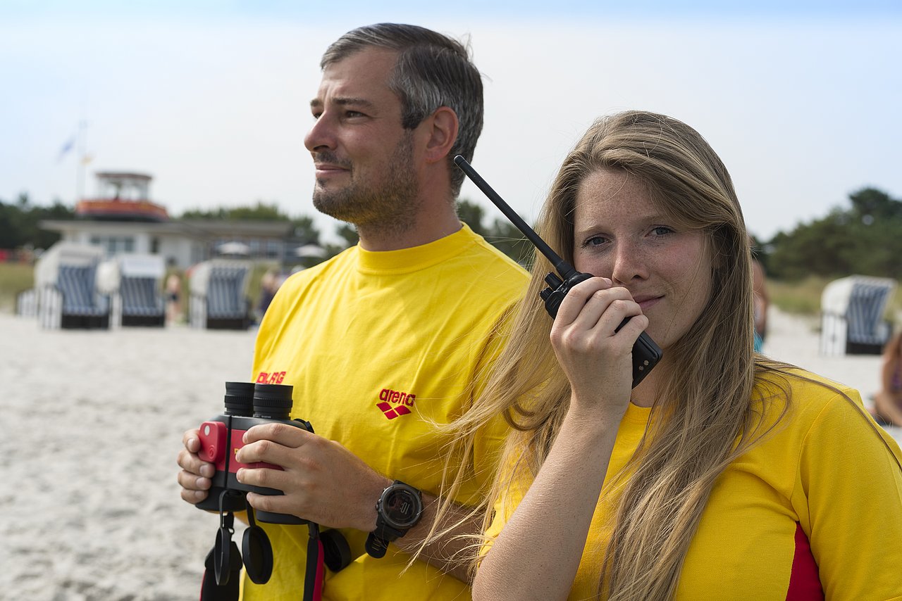 Zwei Mitarbeiter schauen aufs Meer, Frau spricht ins Walkie-Talkie