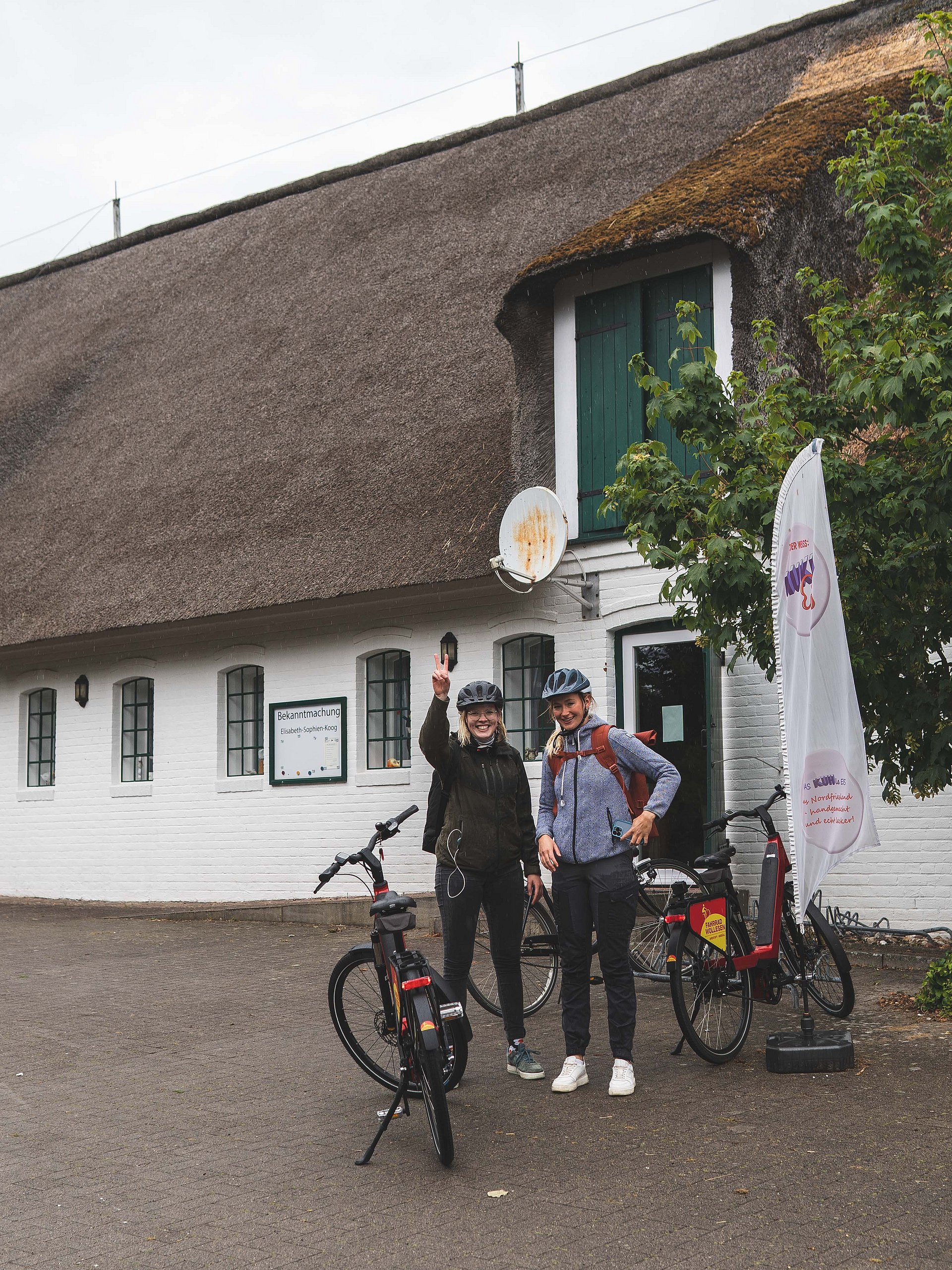 Zwei Radfahrerinnen stehen nach der Hälfte der Radtour über die Insel Nordstrand glücklich und zufrieden vor dem Pharisäerhof Nordstrand. Im Hintergrund ist ein Fachwerkhaus zu erkennen, indem sich der Pharisäerhof befindet. 