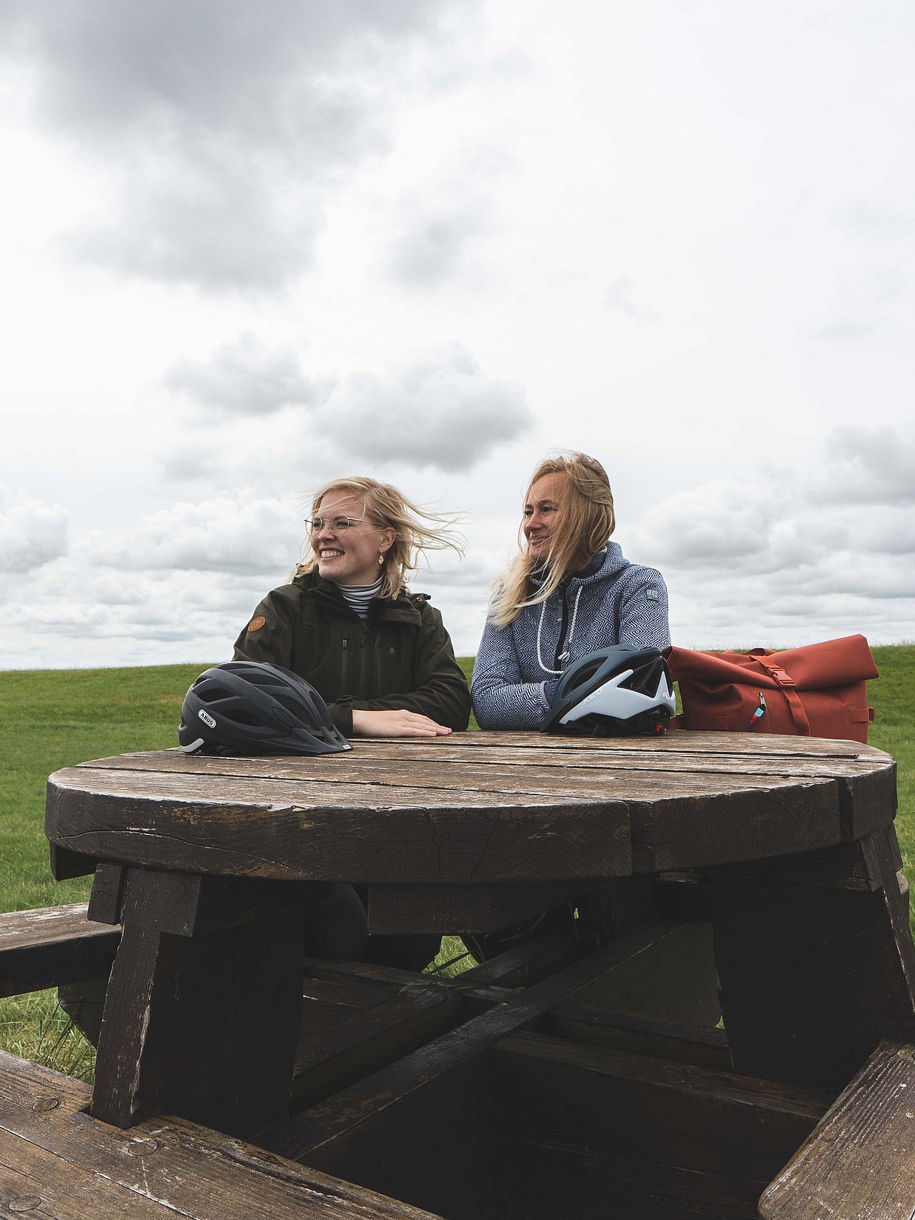 Zwei Radfahrerinnen sitzen an einem Holztisch. Sie blicken beide in die Ferne. Im Hintergrund ist eine Wolkenfront und eine Wiesenlandschaft zu erkennen. 