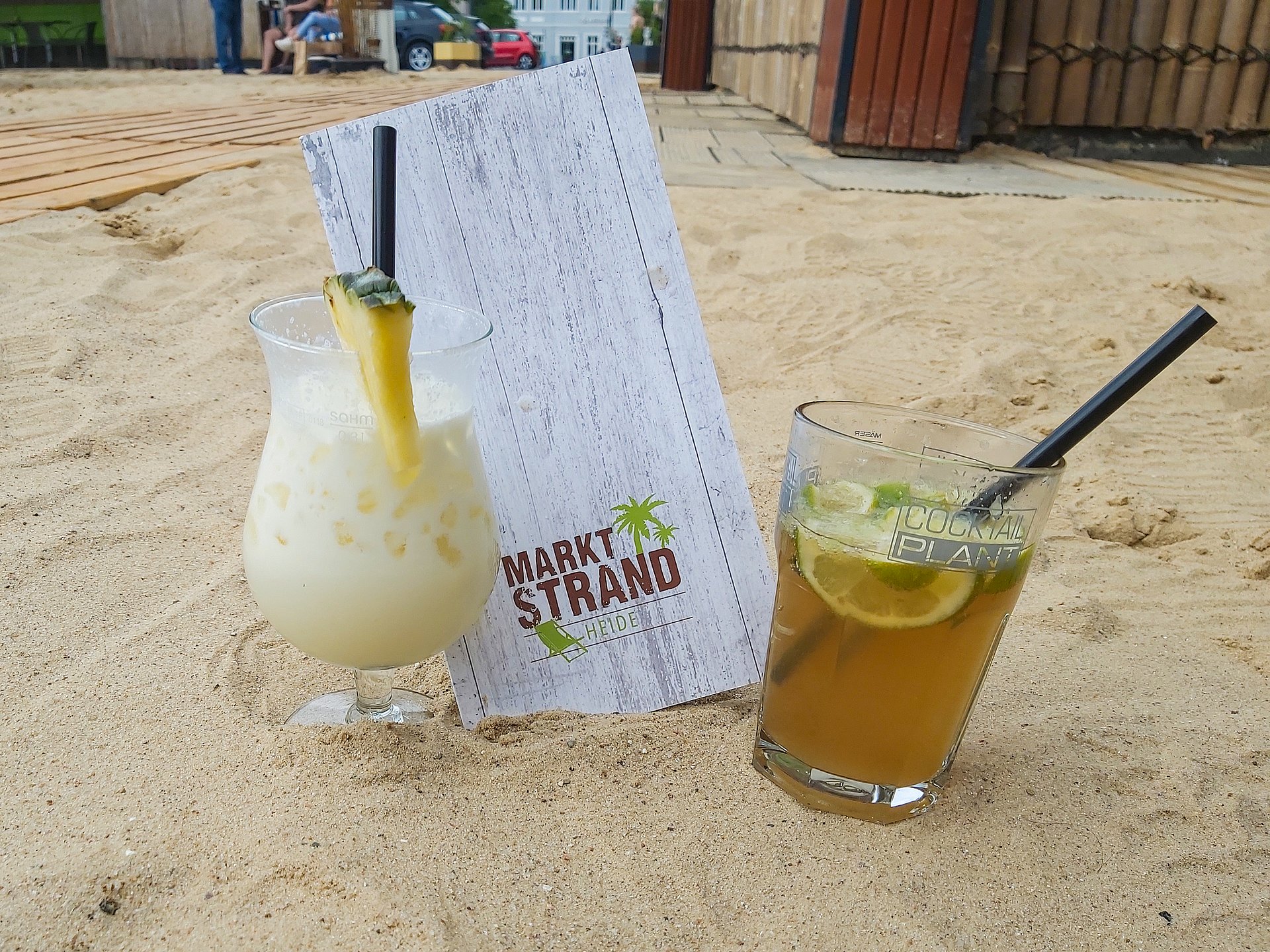 zwei Cocktails und die Broschüre des Marktstrandes stehen im Sand
