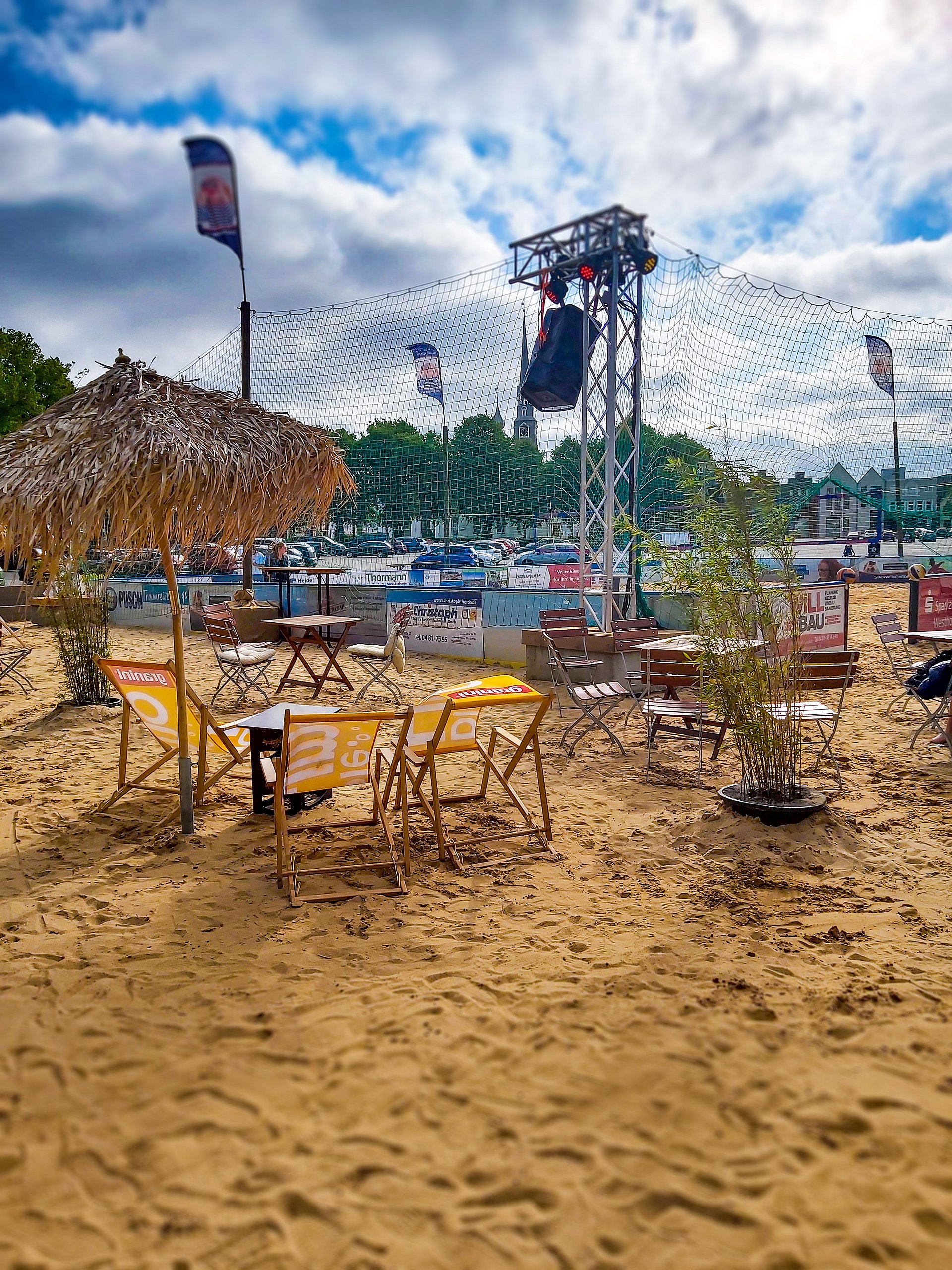 Im Hintergrund ist das Beachvolleyball-Feld zu sehen, im Vordergrund stehen Stühle und Tische im Sand