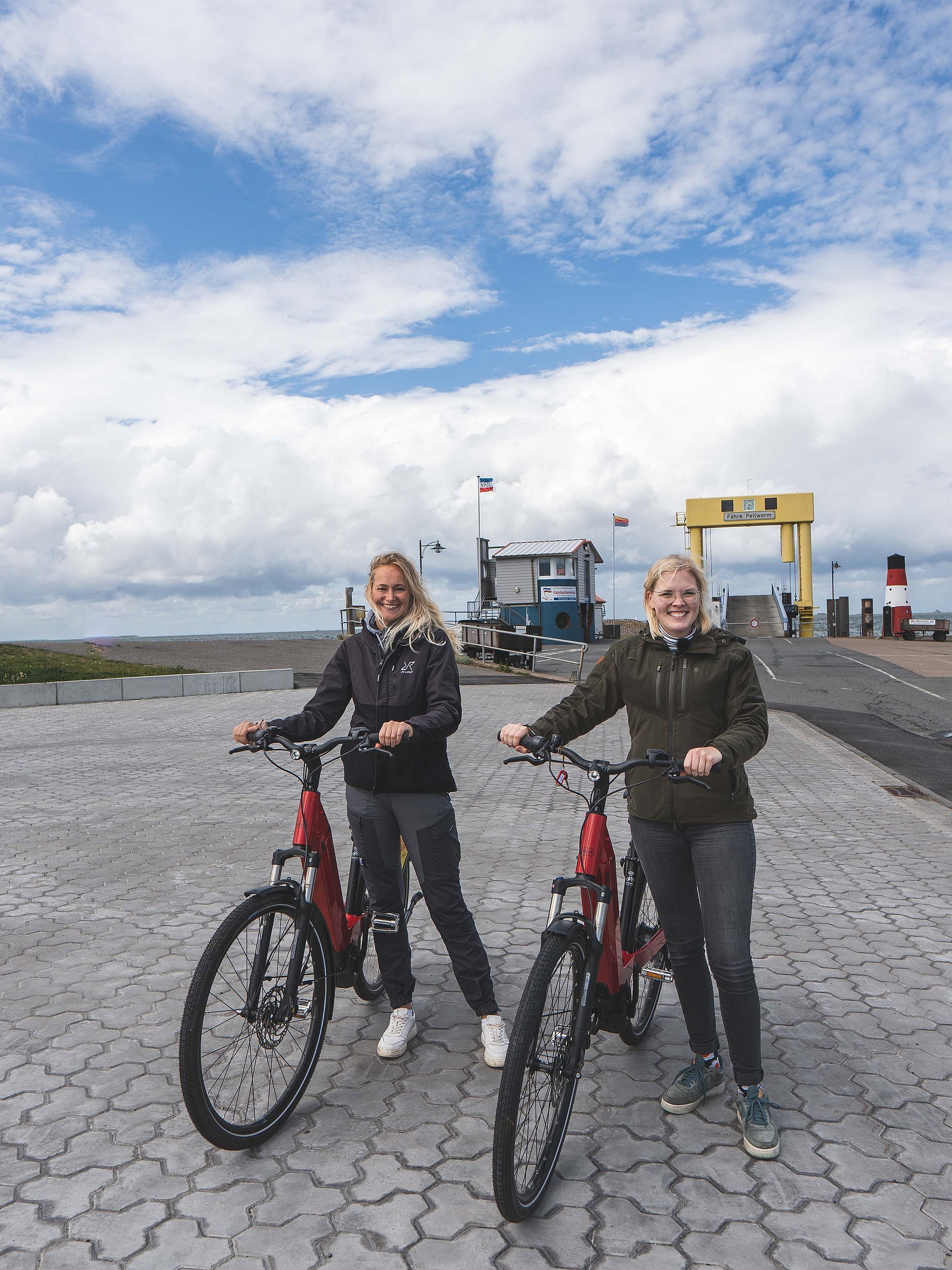 Zwei Radfahrerinnen stehen vor dem Strucklahnungshörn Hafen Nordstrand. Sie haben die Hafenrunde beendet und lächeln glücklich in die Kamara. Im Hintergrund ist ein blauer, aber bewölkter Himmel zu erkennen. 