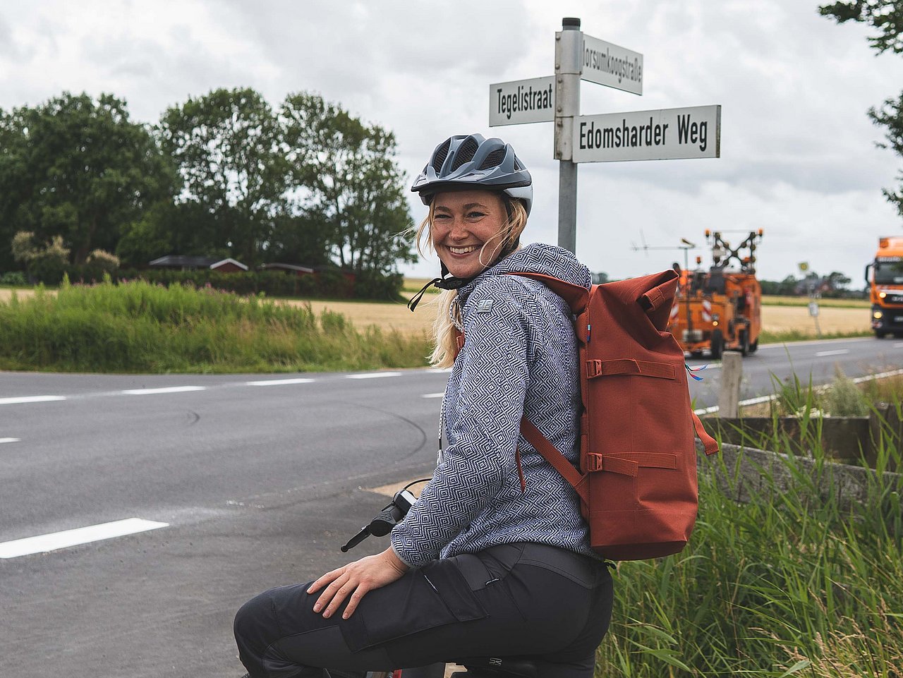 Eine Radfahrerin blickt nach hinten und lächelt. Im Hintergrund sind eine Kreuzung und diverse Straßennamen auf Schildern zu erkennen.