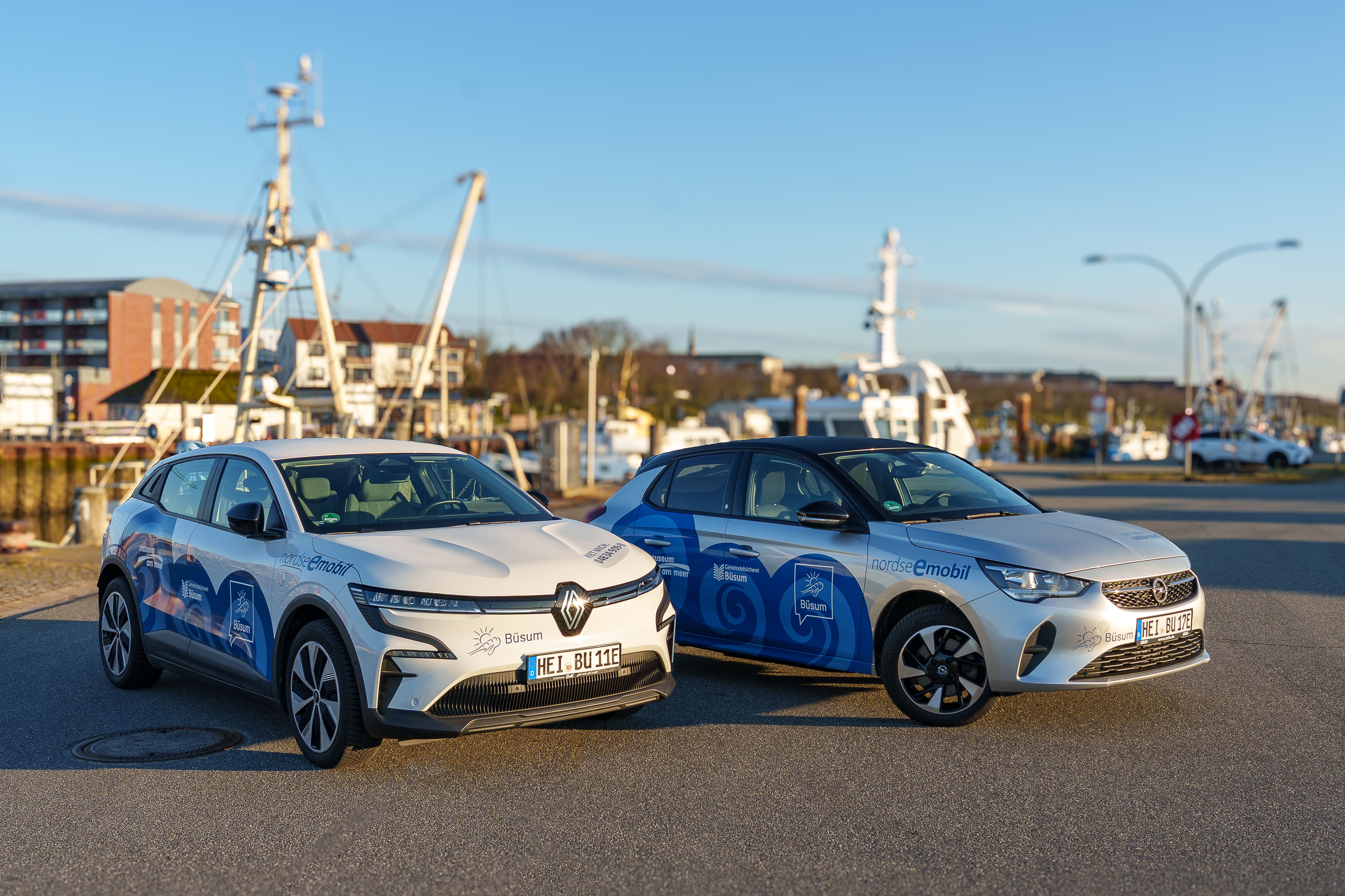 Zwei Nordseemobile (weiße Elektroautos mit blauen Wellendesign) stehen am Museumshafen in Büsum