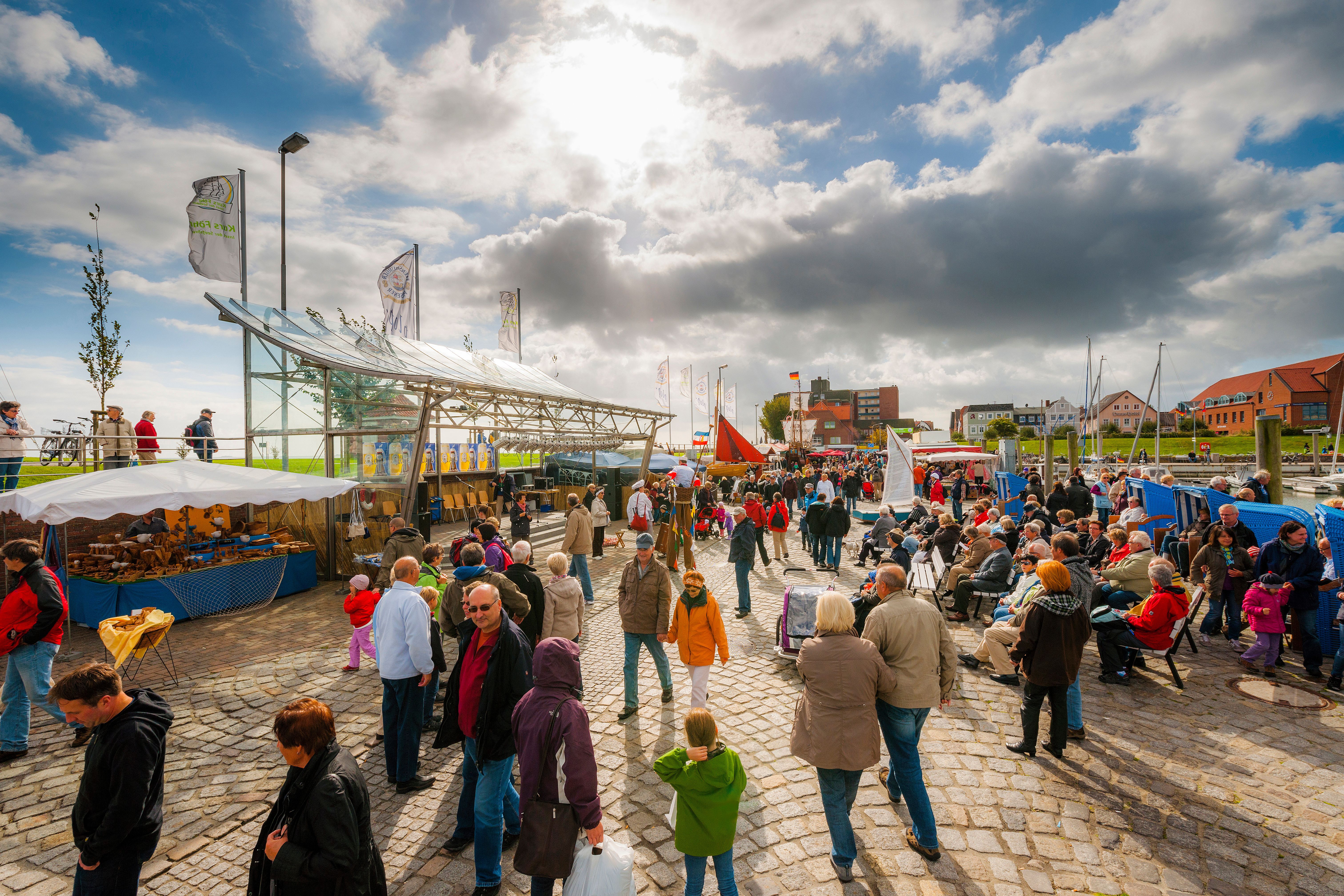 Auf dem Hafenplatz in Wyk auf Föhr herrscht buntes Treiben. Besucher sitzen bereits wartend vor der mit Glas überdachten Bühne, während ander noch auf dem Gelände flanieren. - Foto: Foehr Tourismus GmbH | Oliver Franke
