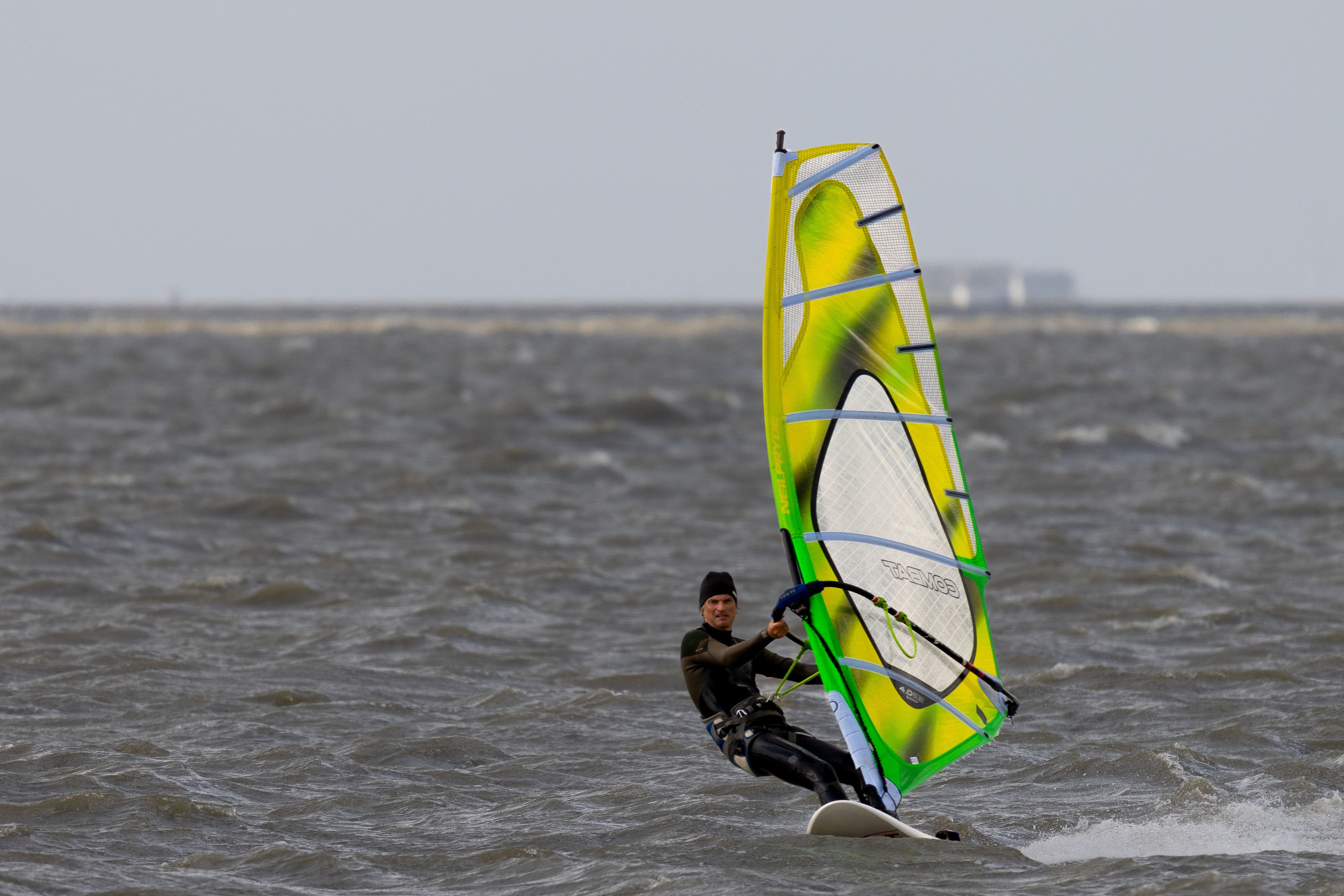 Ein Windsurfer fährt von rechts nach links zügig am Holmersiel auf Nordstrand mit seinem bunten gelb-grünen Segel über die Nordsee. Foto: Gary Asmussen