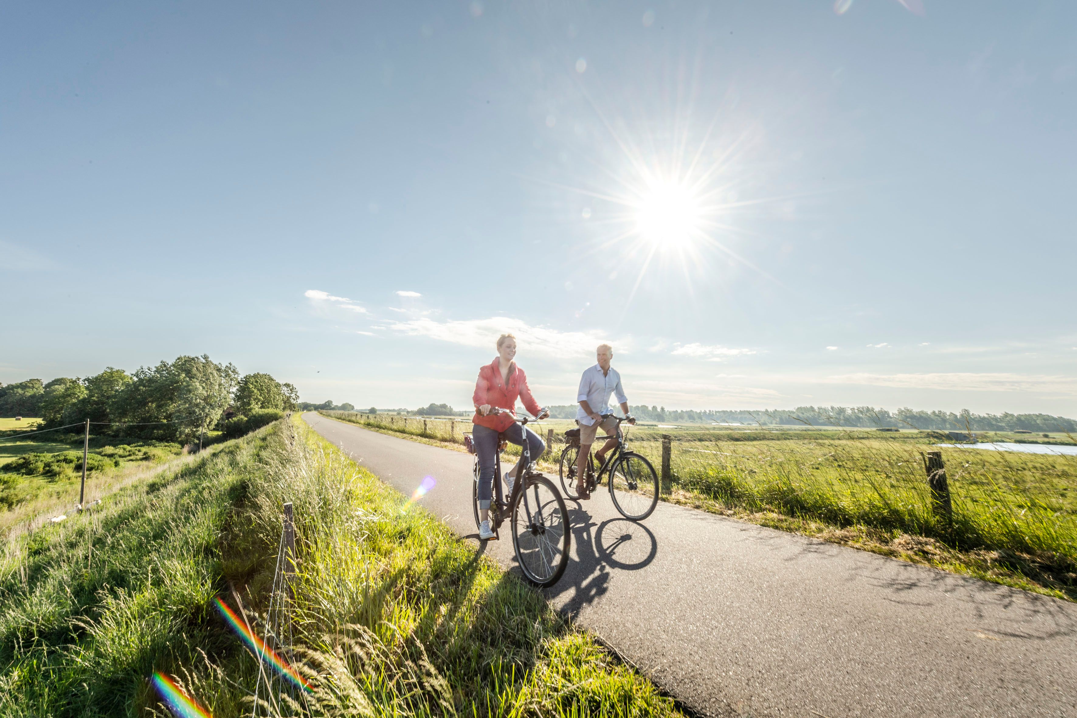 Ein Paar fährt mit Ihren Fahrrädern aus der Sonne kommend auf einem alten Deich, der zur Straße umfunktioniert worden ist.