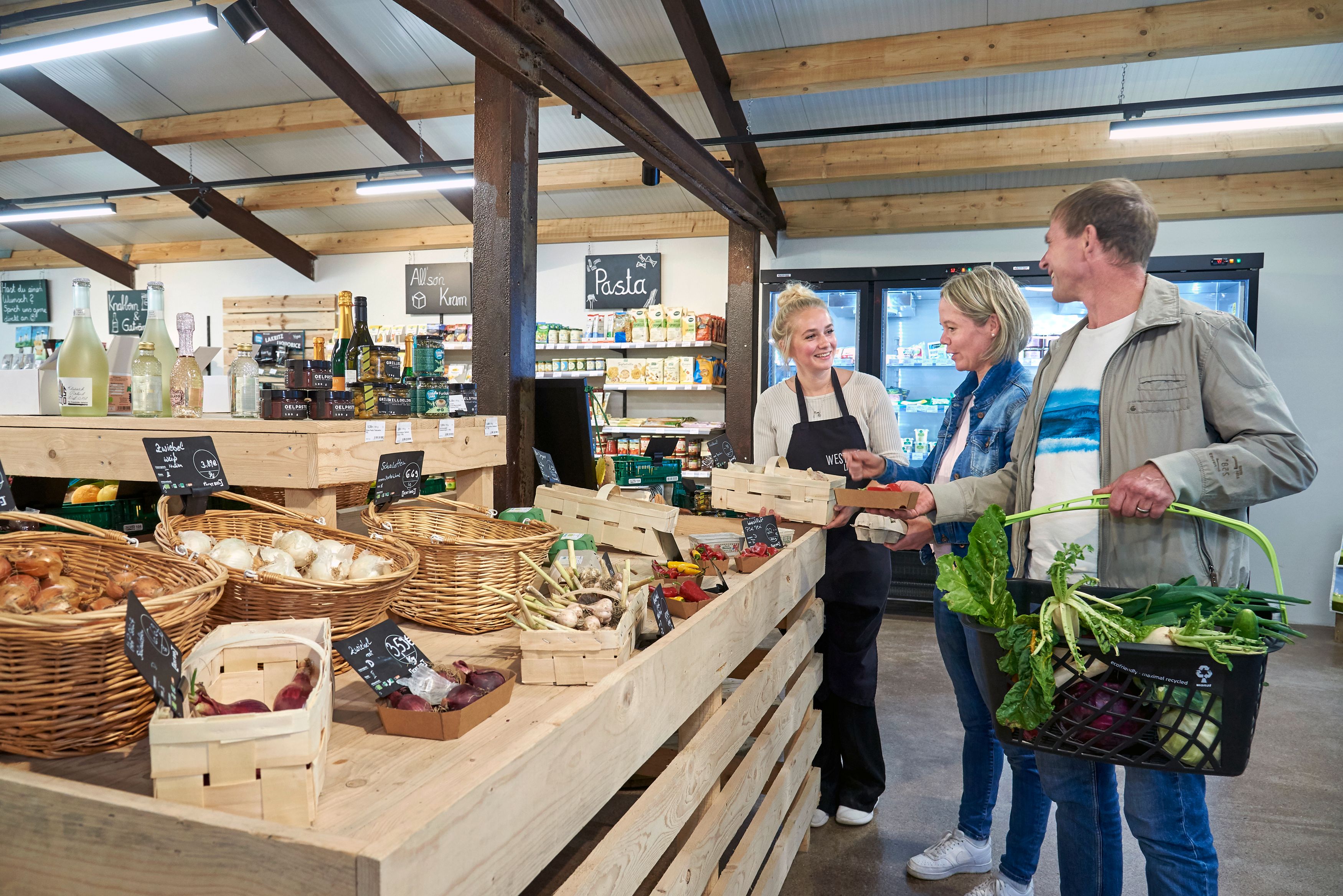 Kunden, eine Frau und ein Mann, stehen in einem Hofladen und lassen sich am Gemüsestand von einer Mitarbeiterin beraten.