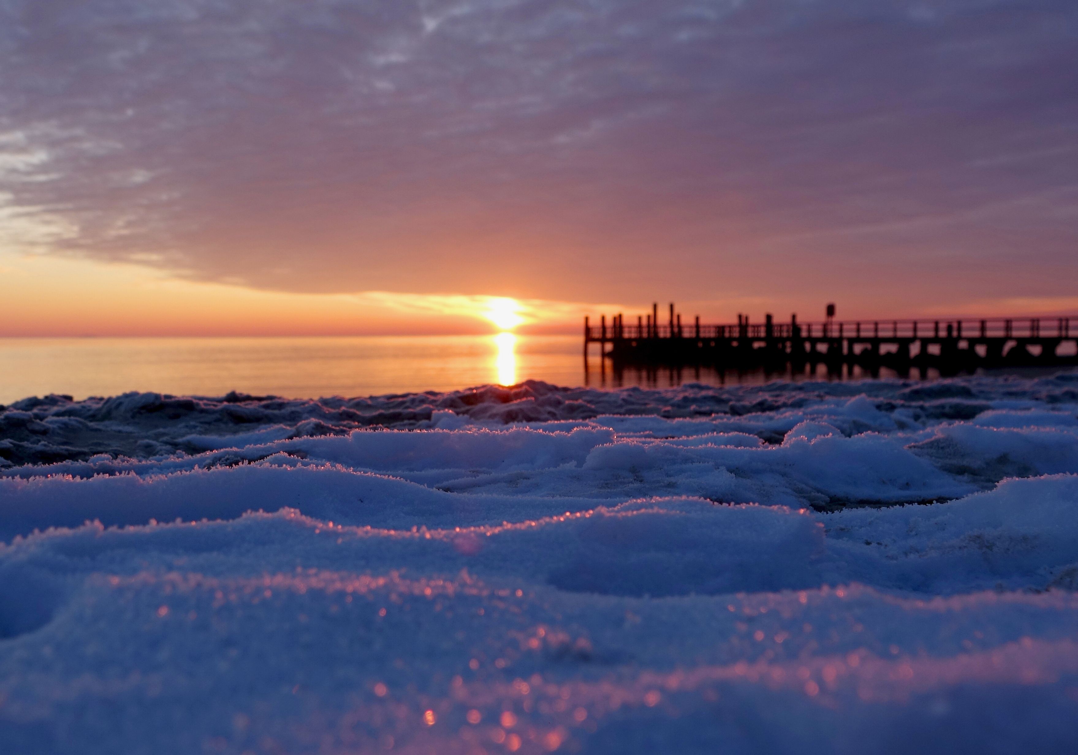 Winterstimmung am Wyker Strand, die Sonne geht über den Meer unter und spiegelt sich im sanft am Strand liegenden Schnee.