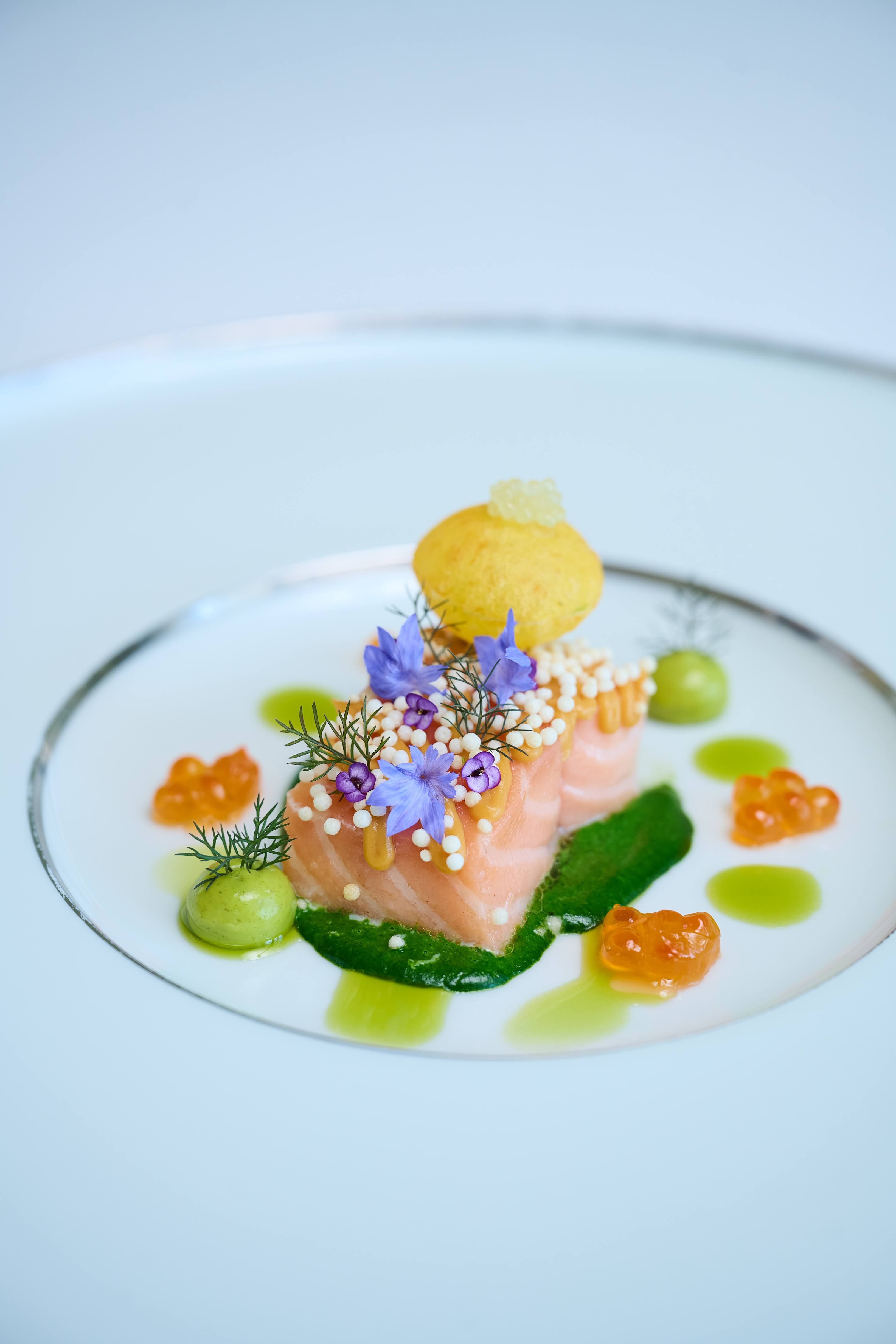 Fischgericht (Lachs) auf einem weißen flachen Teller mit zwei silbernen, weitauseinanderliegenden dünnen Kreisen. Foto: GH Heiligendamm