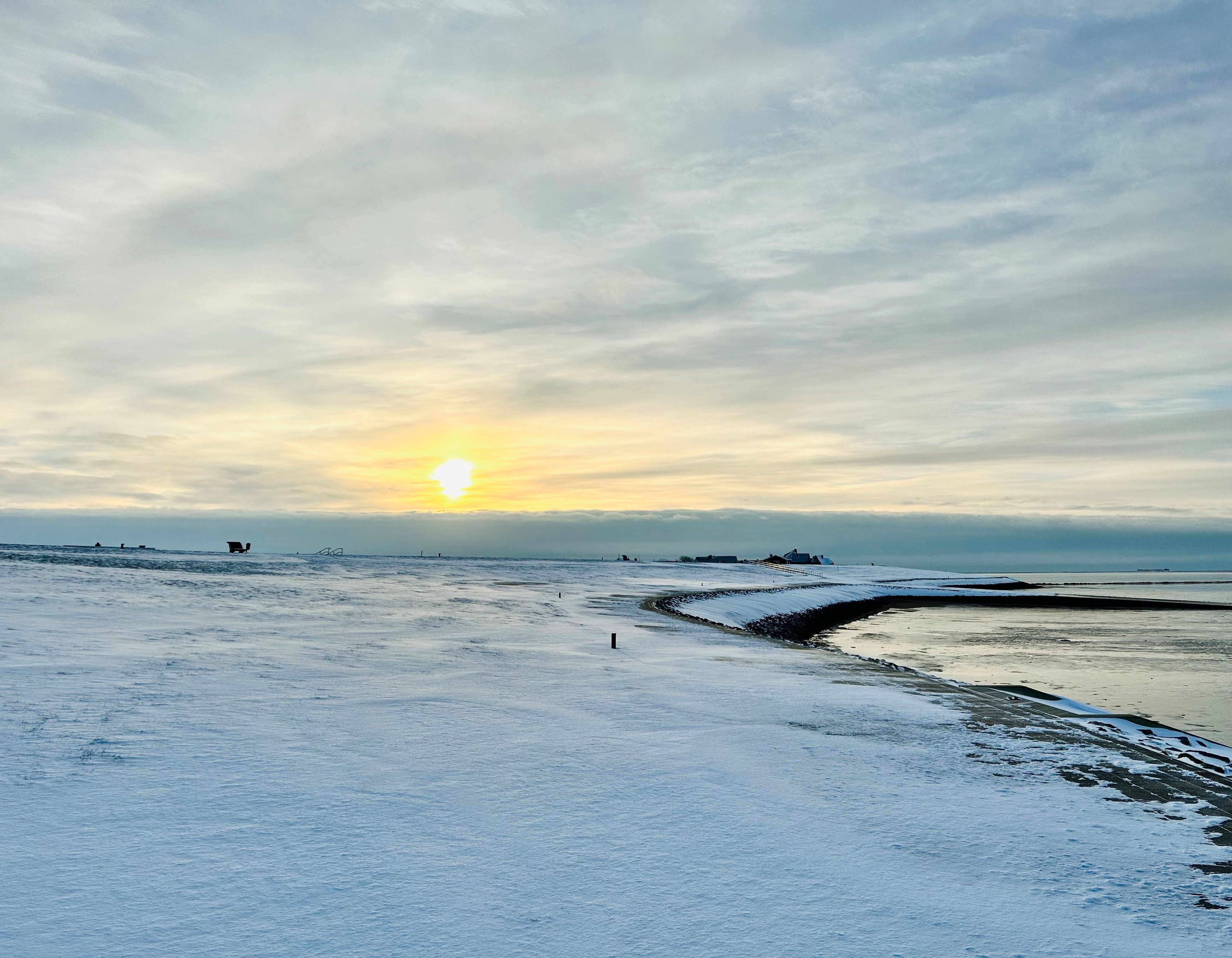 Verschneiter Deich von Dagebüll bei tiefstehender Sonne. Es ist ein windstiller Tag, der Schnee liegt sanft da. 