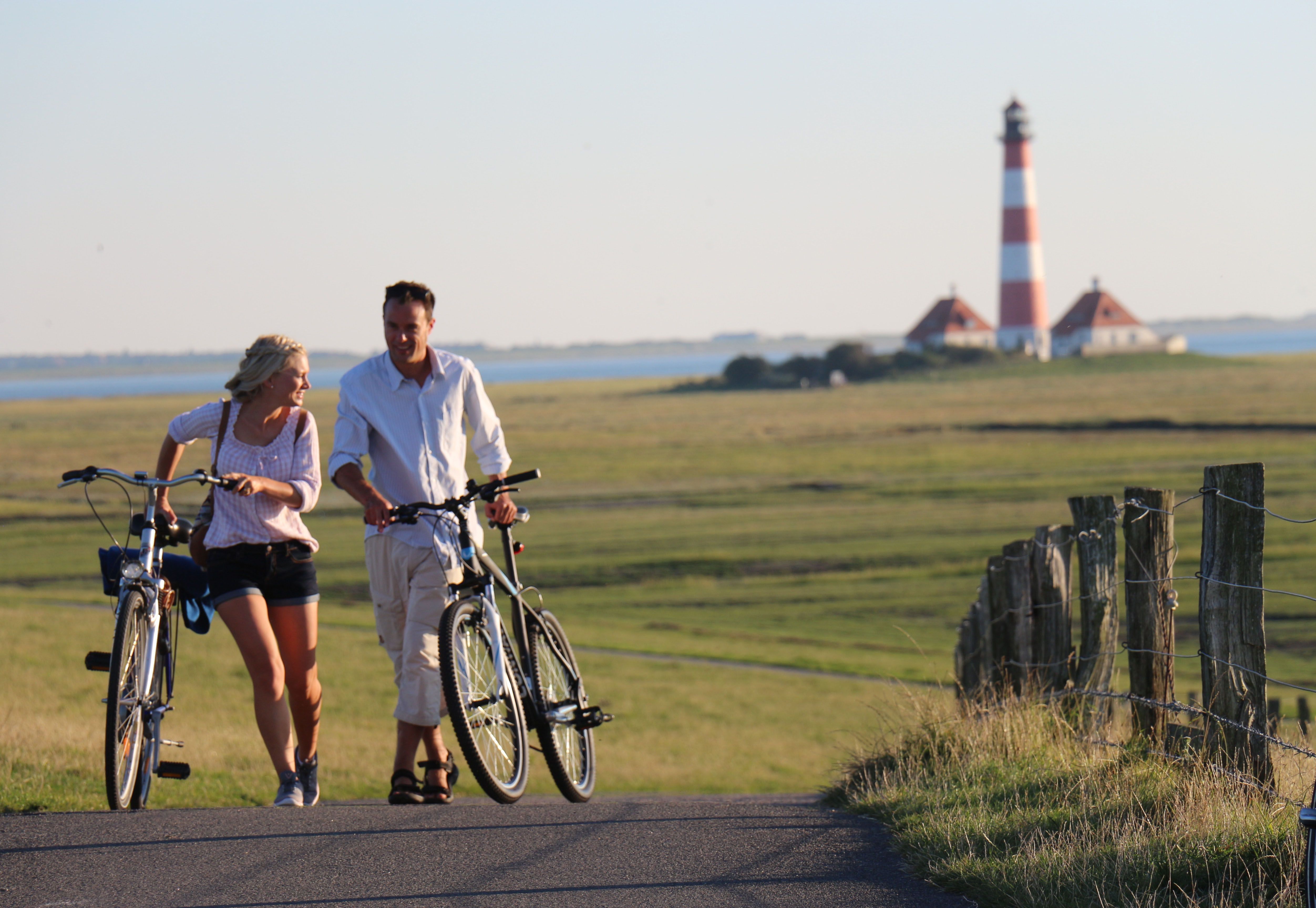 Ein Paar schiebt seine Fahrräder die Deichauffahrt nach einem Besuch am Leuchtturm Westerheversand hoch. Dabei ist der Leuchttrum mit seinen beiden markanten Leutchtturmwärterhäusern im Hintergrund zusehen.