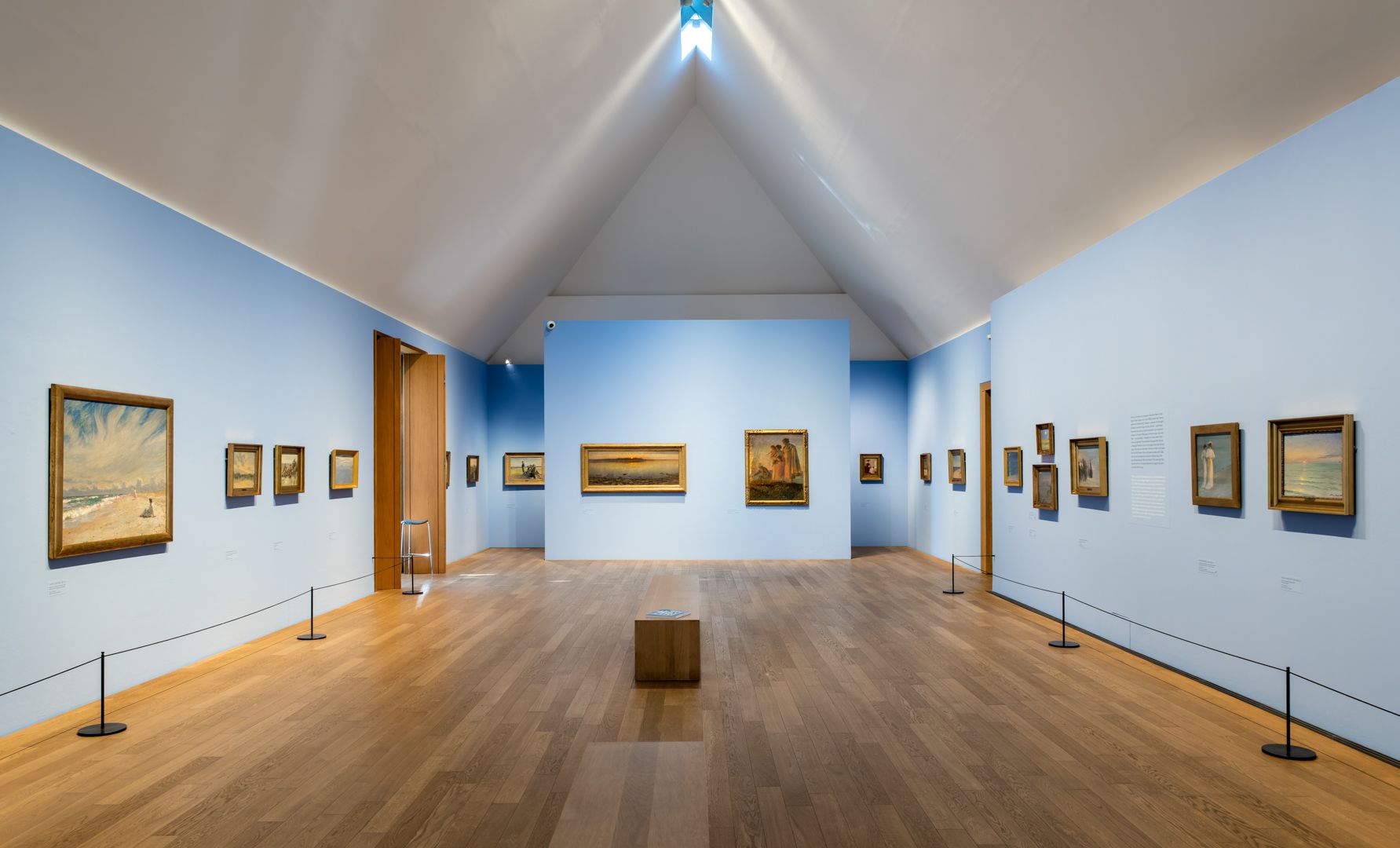 Ein Ausstelllungsraum des Museums Kunst der Westküste, in einer Weitwinkelaufnahme. Zu sehen sind die linke, die rechte sowie die hintere Wand mit Bildern und der freie Raum mit einer Sitzgelegenheit zum Verweilen. Die Wände sind hellblau und mit Bildern behängt. Der Boden ist mit hellem Pakett belegt. 