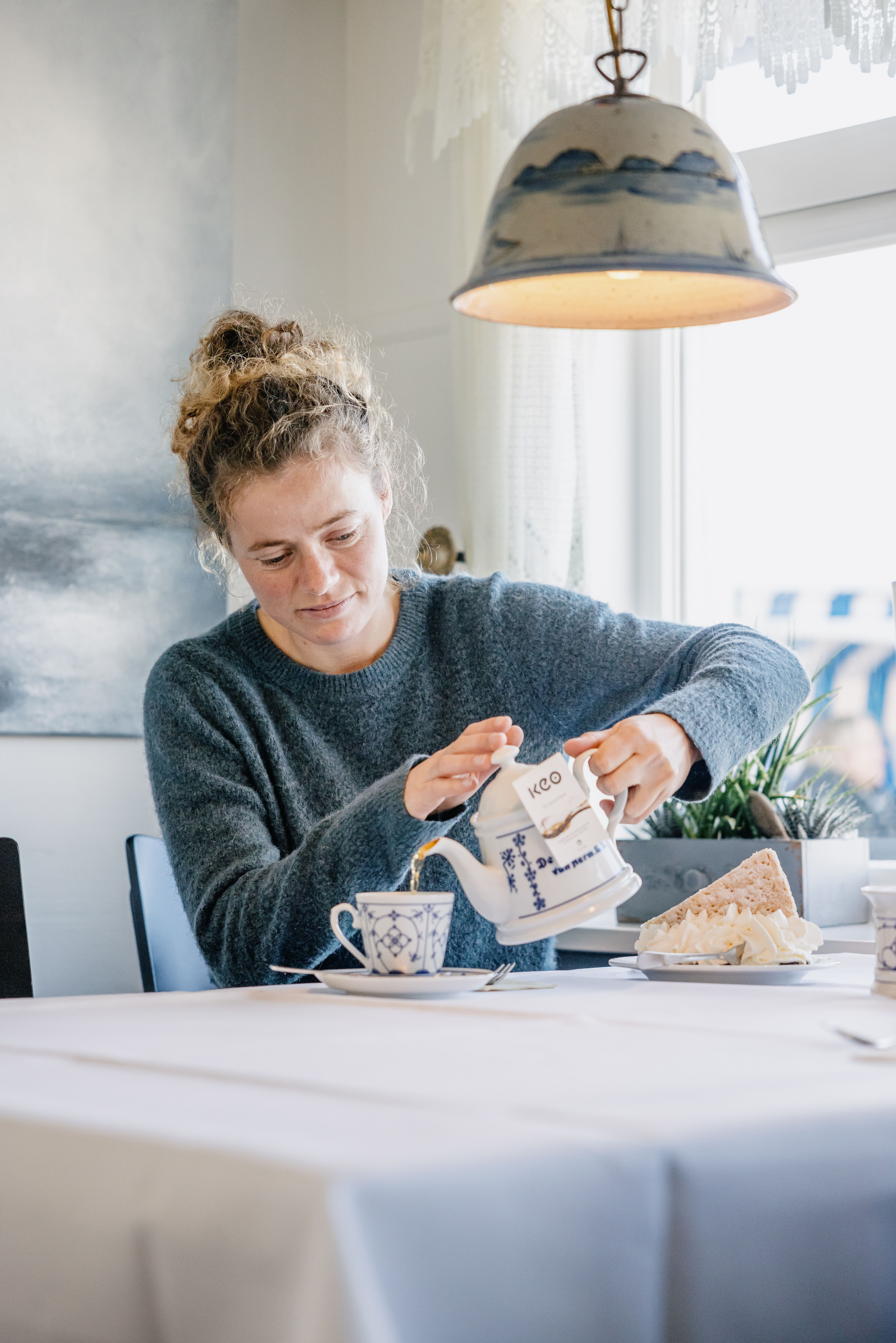 Eine Frau sitz an einem mit einer weißen Tischdecke dedekchten Tisch und schenkt sich aus einer Kanne im Friesendesign Tee in ihre Friesentasse.