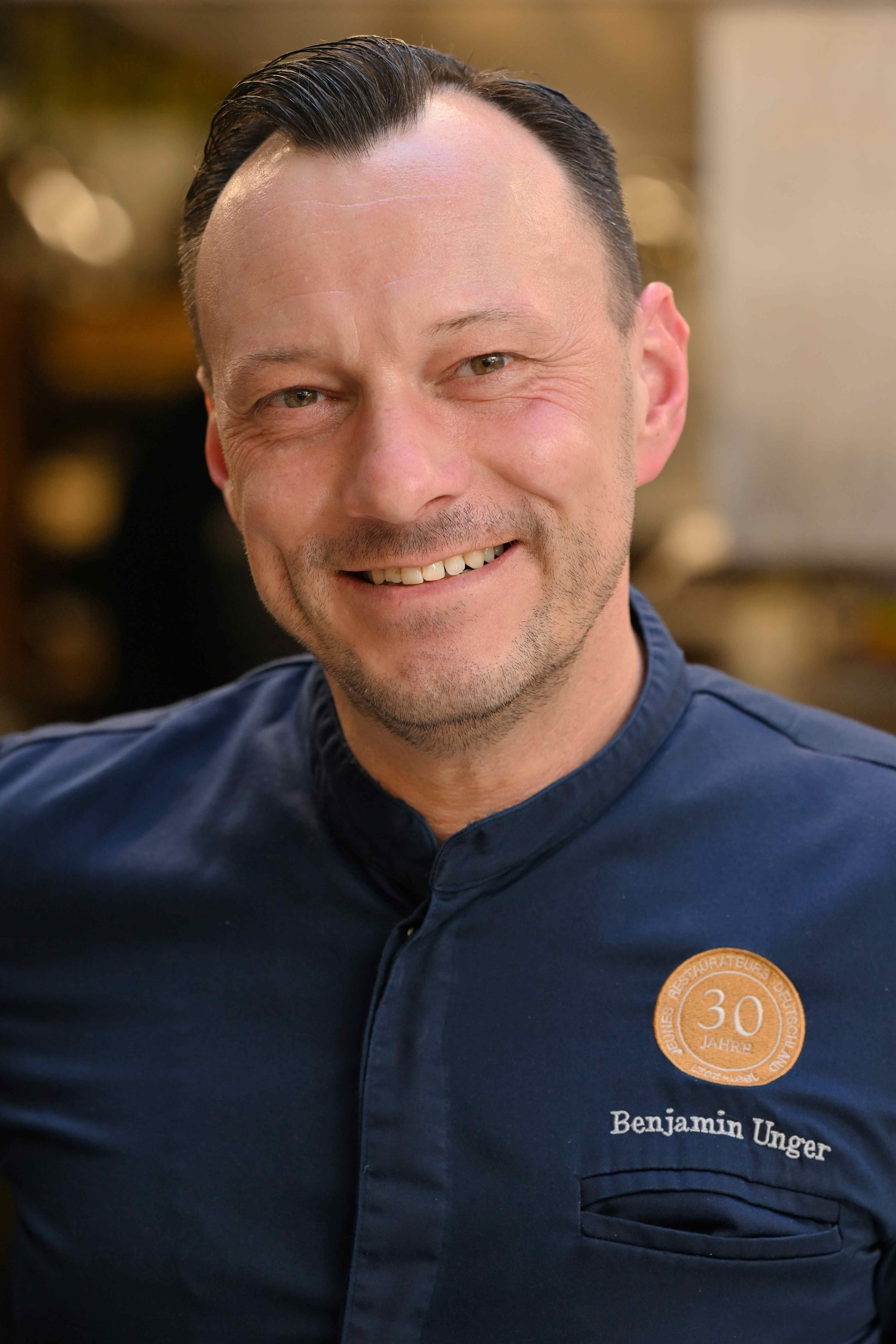 Benjamin Unger lächeld in dunkelblauer Kochjacke mit Benjamin Unger Logo in die Kamera. Foto B. Unger