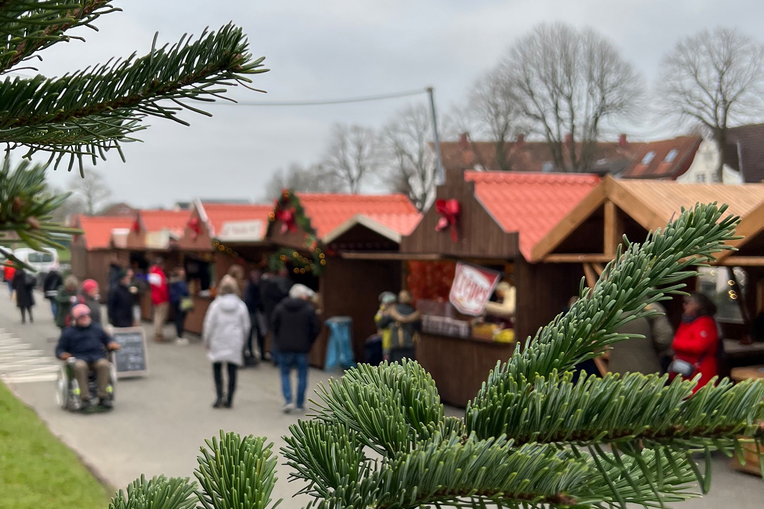 Weihnachtshütten stehen an einem Weg nebeneinander. Passanten spazieren bei mildem Winterwetter daran vorbei oder stehen davor.