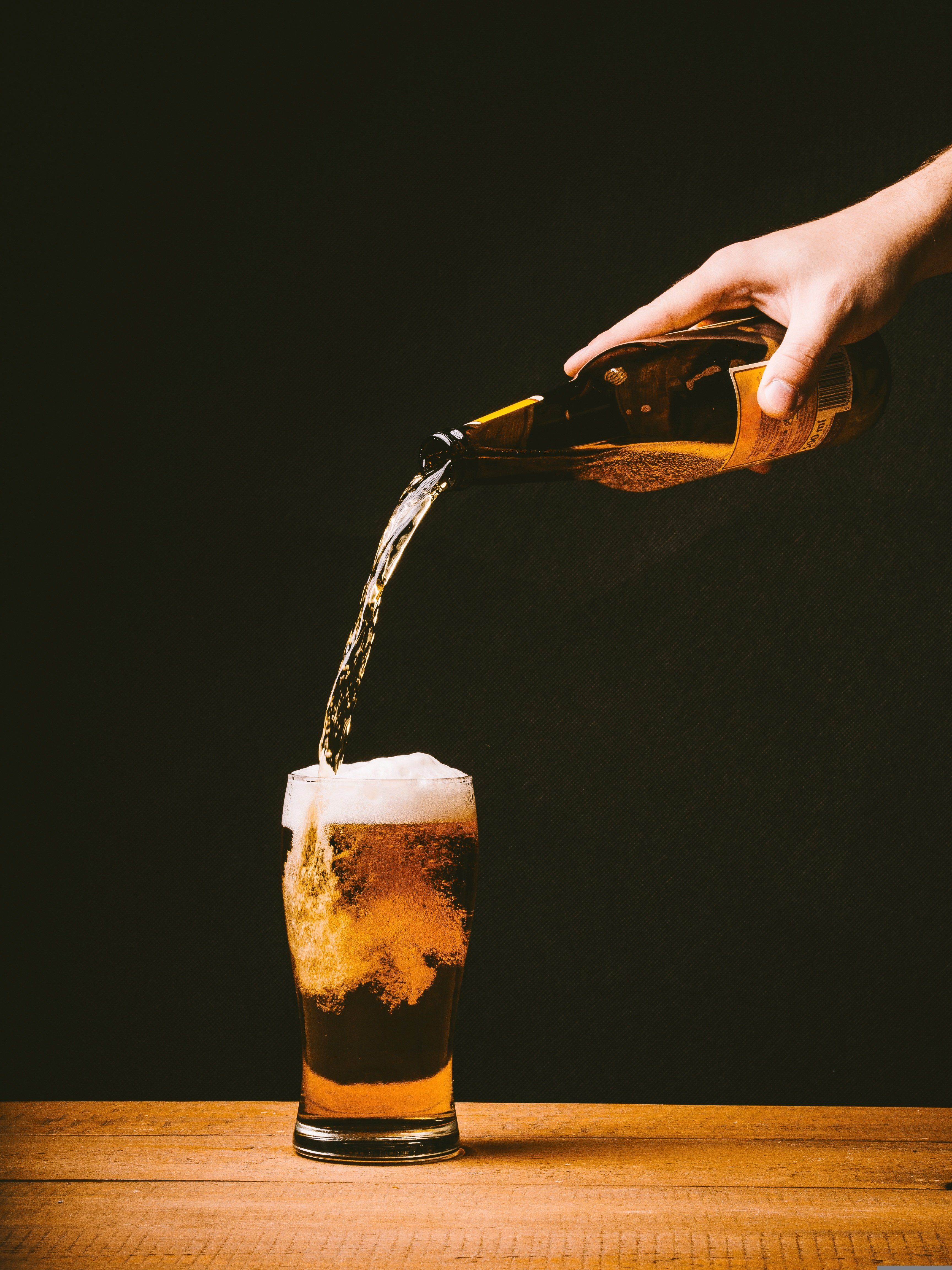 Jemand schenkt vor dunklem Hintergrund aus einer Flasche im hohen Bogen Bier in ein auf einem Holztisch stehendes Bierglas. Foto:  Republica - pixabay