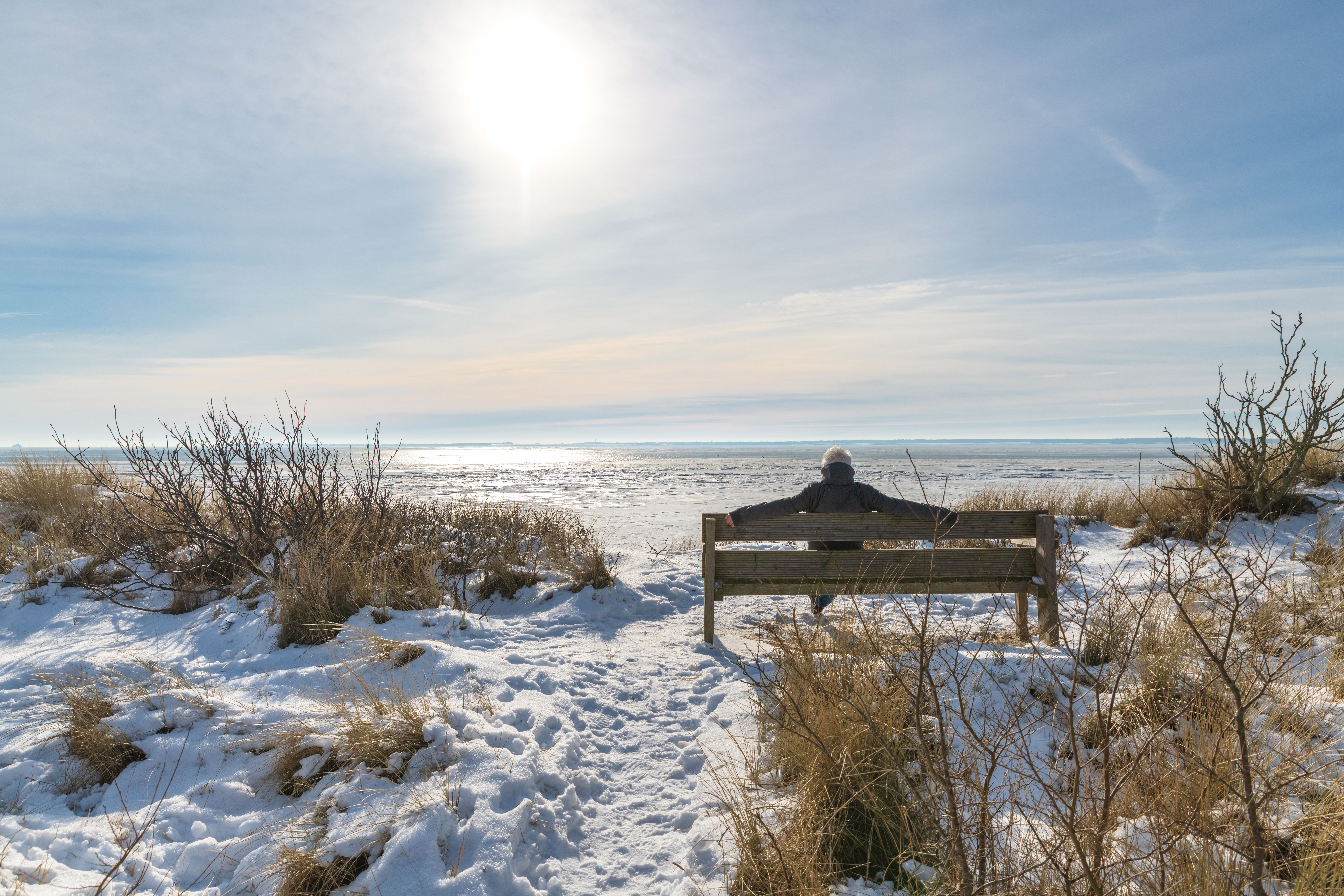 Ein Mann sitz im Winter mit ausgebreiteten Armen auf einer schneebedeckten Düne am Strand vom Gotin Kliff mit Blick auf das am Horizont sichtbare Amrum. 