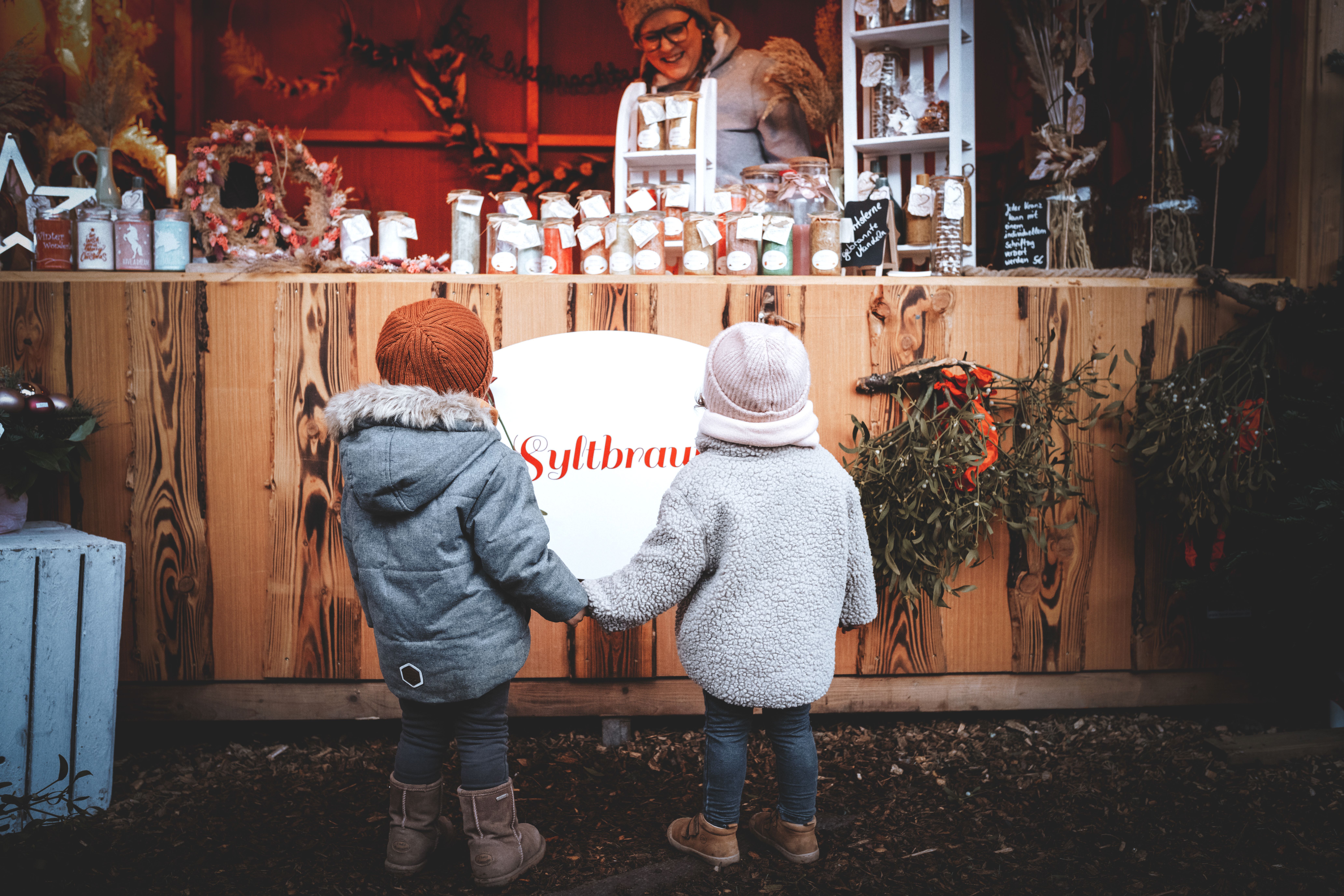 Zwei kleine Kinder stehen in Winterbekleidung mit Mützen Hand in Hand vor eine Weihnachstmarktbude an der handgemachte Kerzen verkauft werden. Die Verkäuferin unterhält sich mit den beiden.
