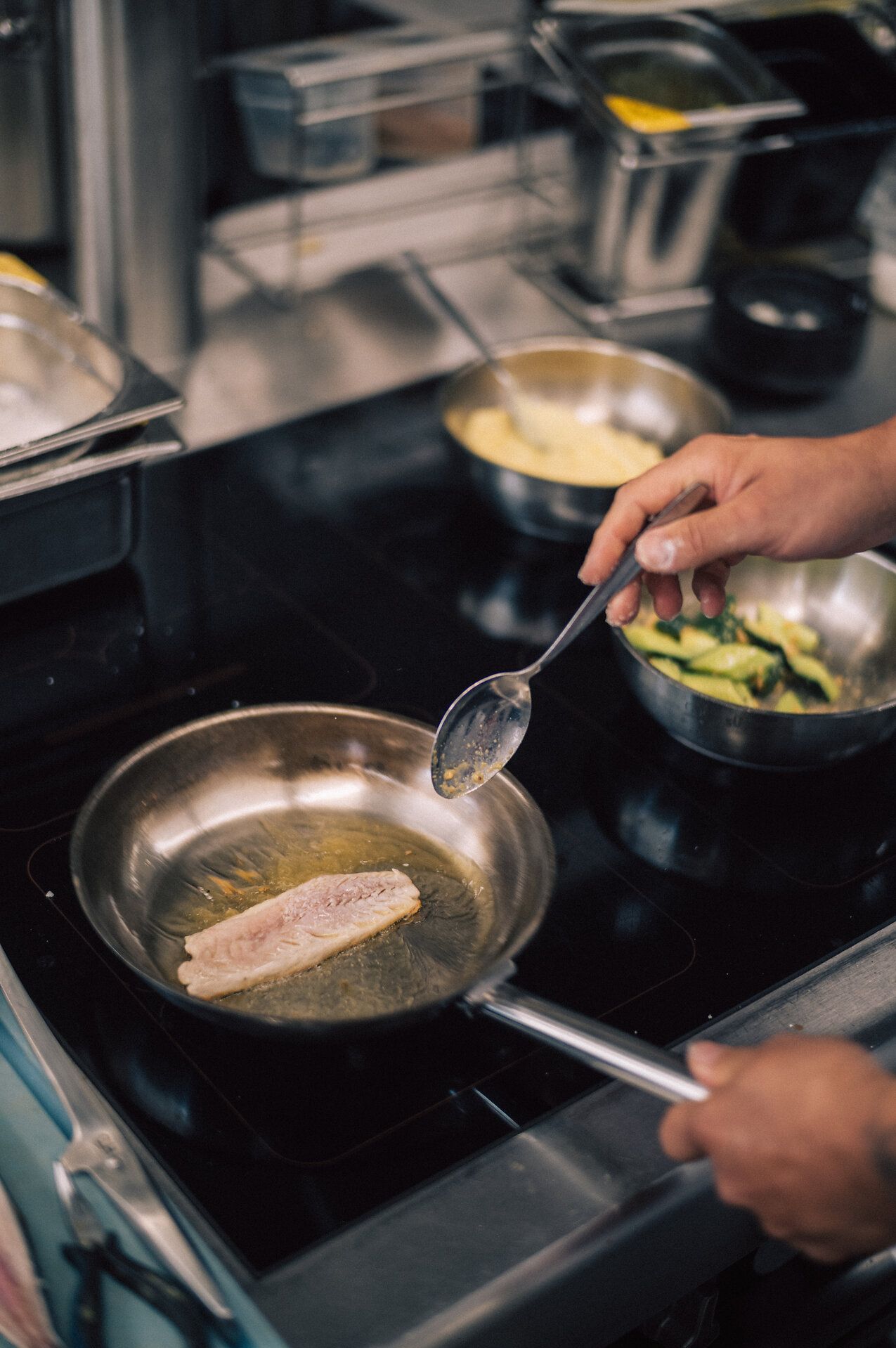Man sieht eine Pfanne in dem ein Stück Fischfilet brät. Ein Koch, es sind nur seine Hände zusehen, hält dabei die Pfanne, während seine rechte Hand mit einem Löffel das Filetstück mit der Butter in der der Fisch brät übergießt.