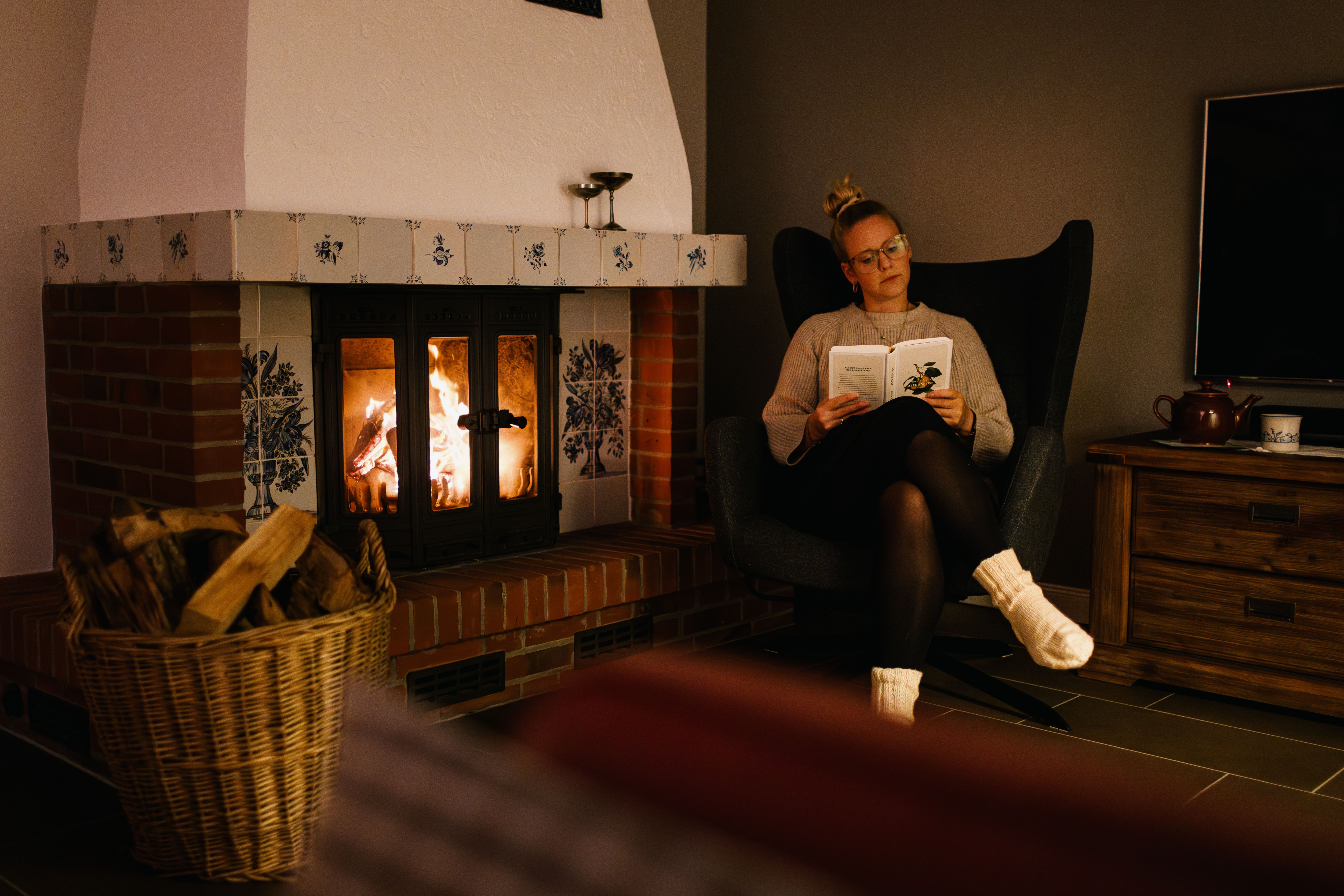 Eine Frau sitzt gemütlich mit einem Buch vor dem brennenden Kamin im Ferienhaus in Nordfriesland.