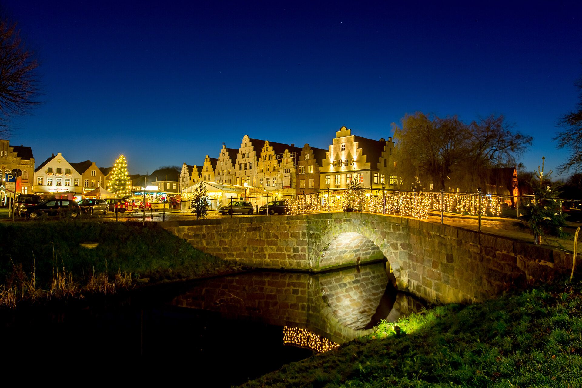 Der Blick geht über die große Gracht und Brücke auf den am Abend weihnahtlich beleuchtetet Friedrichstädter Marktplatz mit seinen holländischen Giebelhäusern.