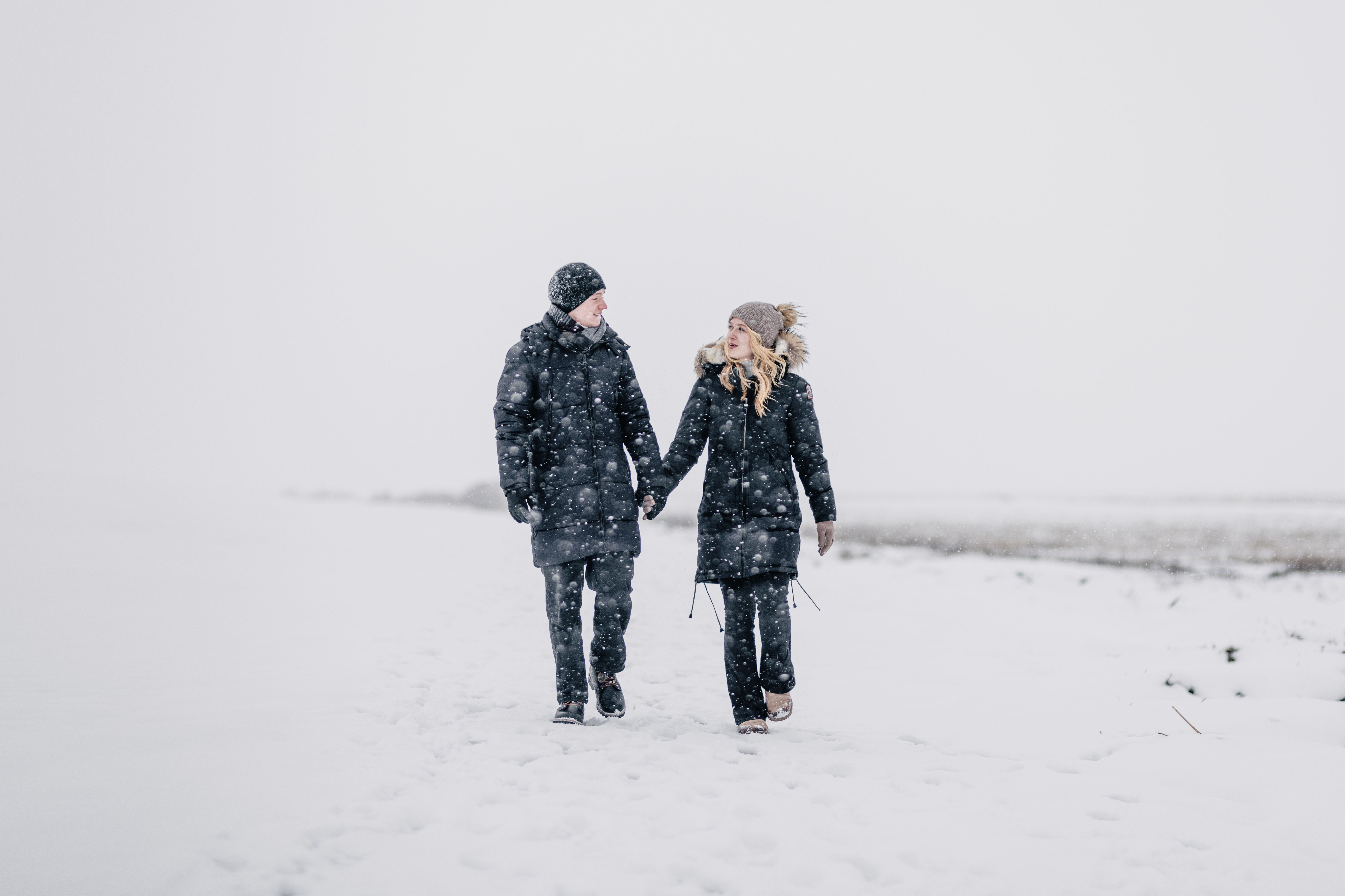 Ein Paar geht, warm eingekleidet mit Handschuhen, Mütze und Schaal, Hand in Hand bei echtem nordfriesischem Winterwetter im Vorland spazieren. Es schneit und außer einem Schilfbereich im rechten Hintergrund ist vor lauter Weiß nicht viel von der Landschaft zu sehen.