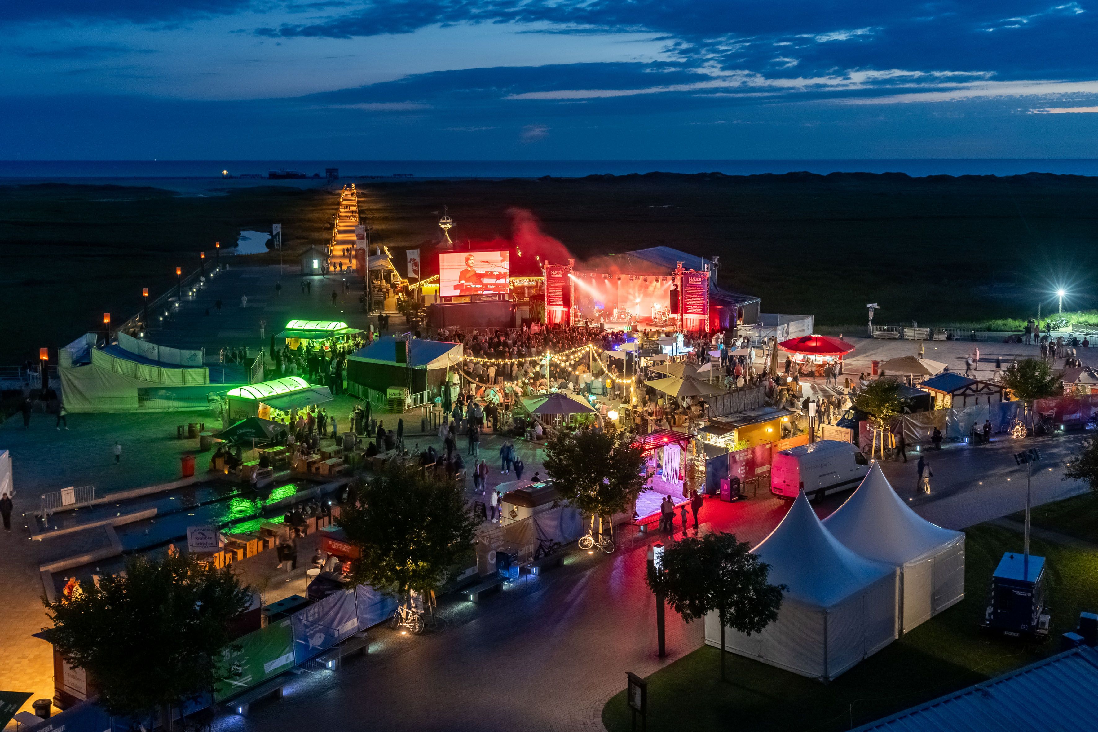 Das Festivalgelände aus der Luft bei Nacht. Bunte Lichter der Bühne und der Buden verzaubern die Betrachtenden