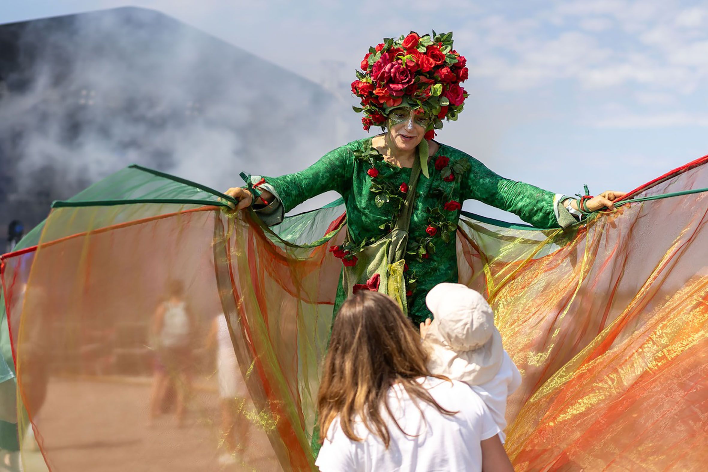 Ein soganannter Walking Act geht auf Stelzen mit bunten Schwingen über das Festivalgelände.