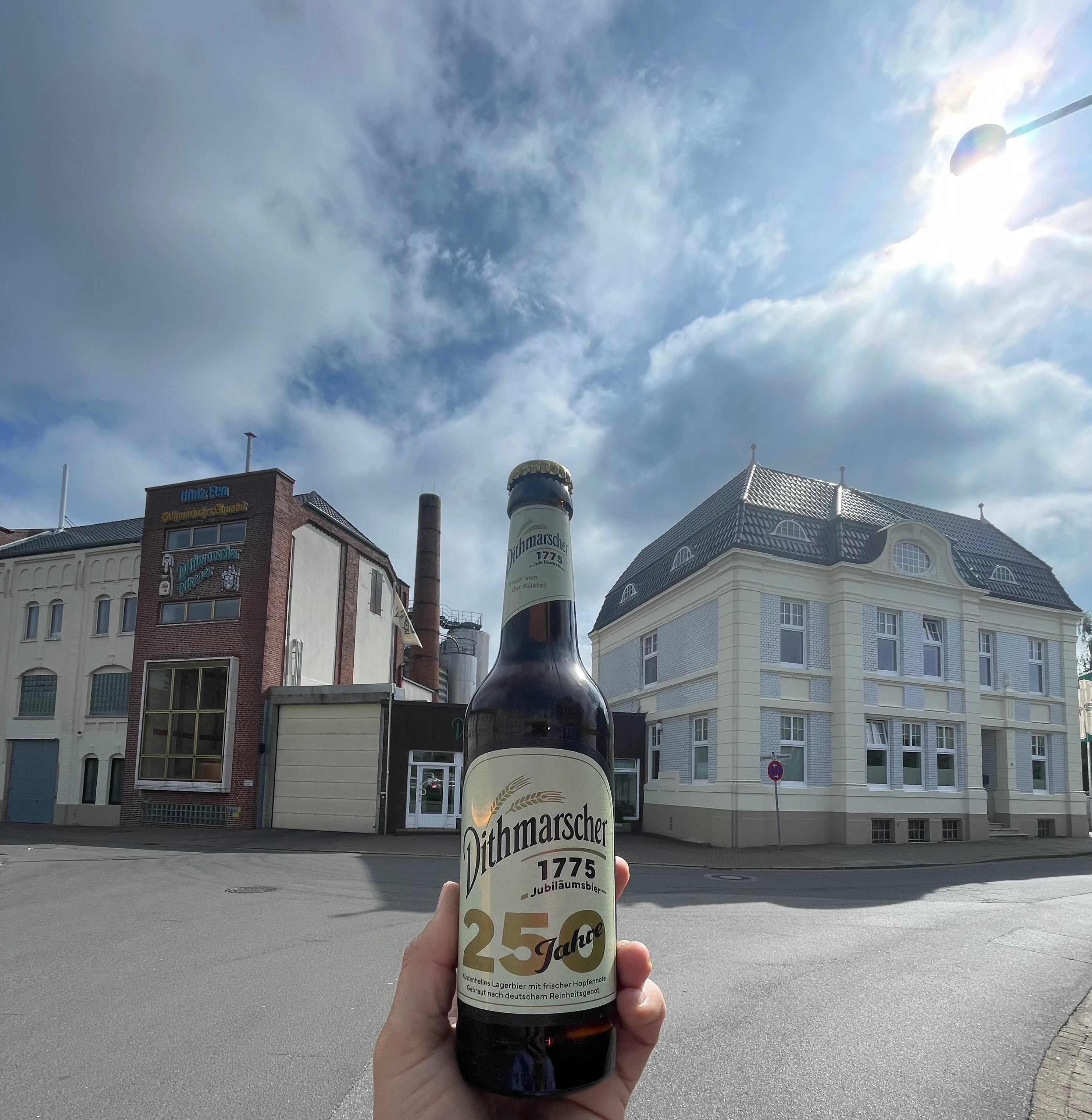 Eine Flasche Bier wird in die Kamera gehalten, im Hintergrund sind Häuser zu sehen