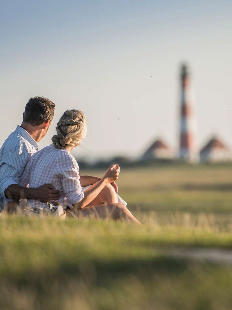 Ein Pärchen sitzt im Gras eines Deiches auf Eiderstedt und blickt auf den Westerhever Leuchtturm
