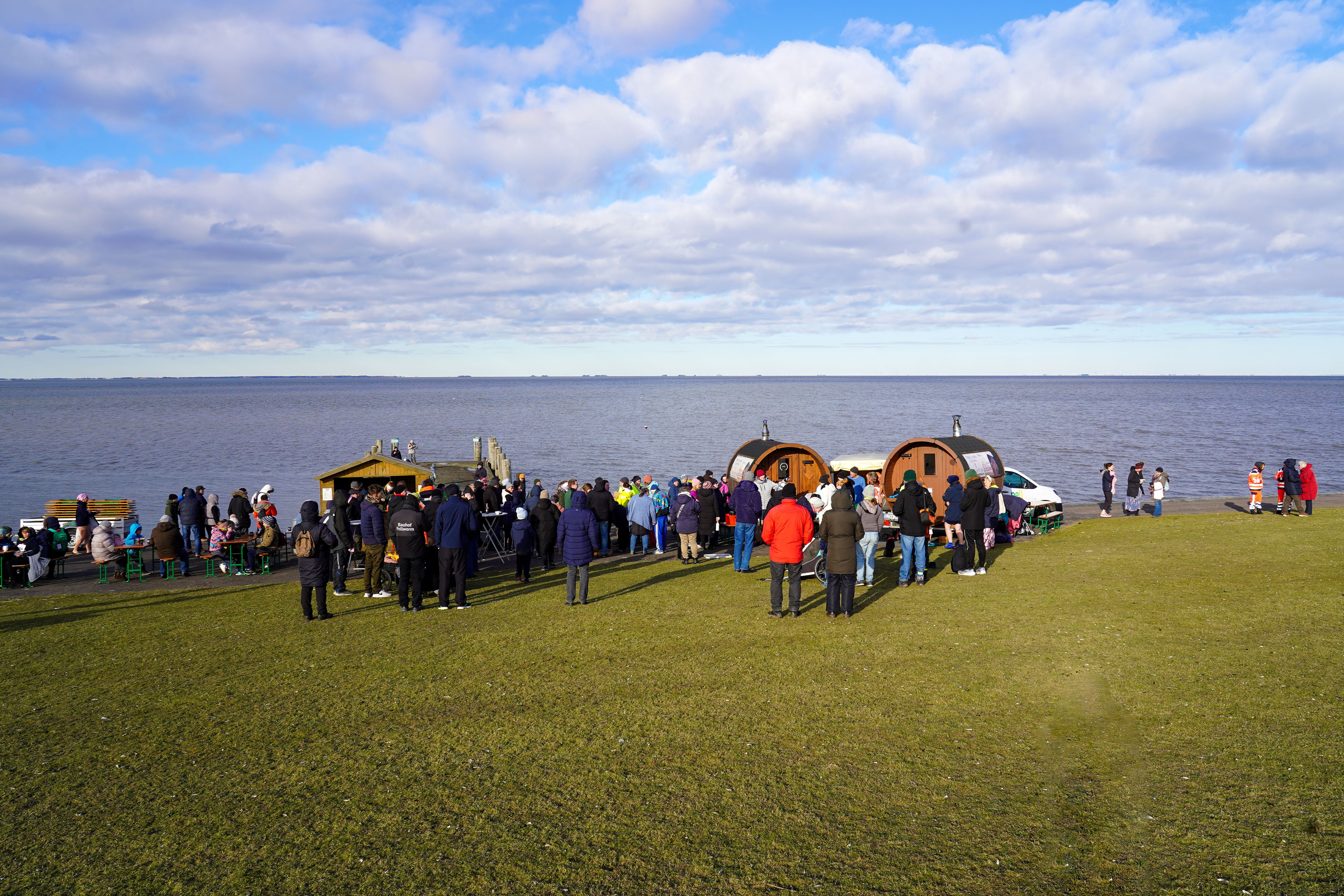 Eine Gruppe von Menschen versammelt sich an einem Ufer mit Verkaufsständen. Der Himmel ist bewölkt, und das Meer erstreckt sich im Hintergrund.