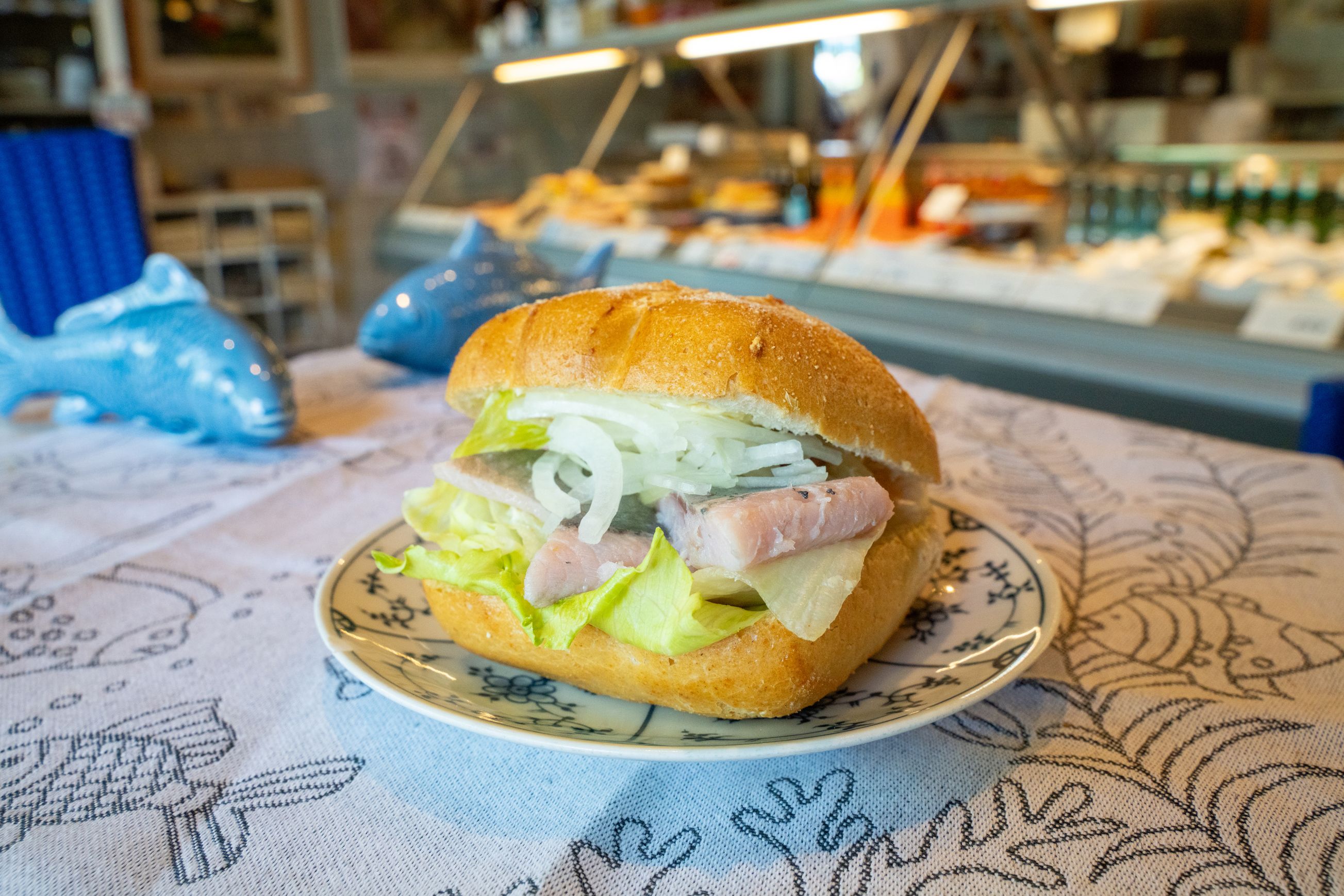 Ein knackfrisches leckeres Fischbrötchen mit Zwiebeln und Salat, präsentiert auf einem typischen Teller mit Friesendesign. - Foto: Föhr Tourismus GmbH/Moritz Kertzscher