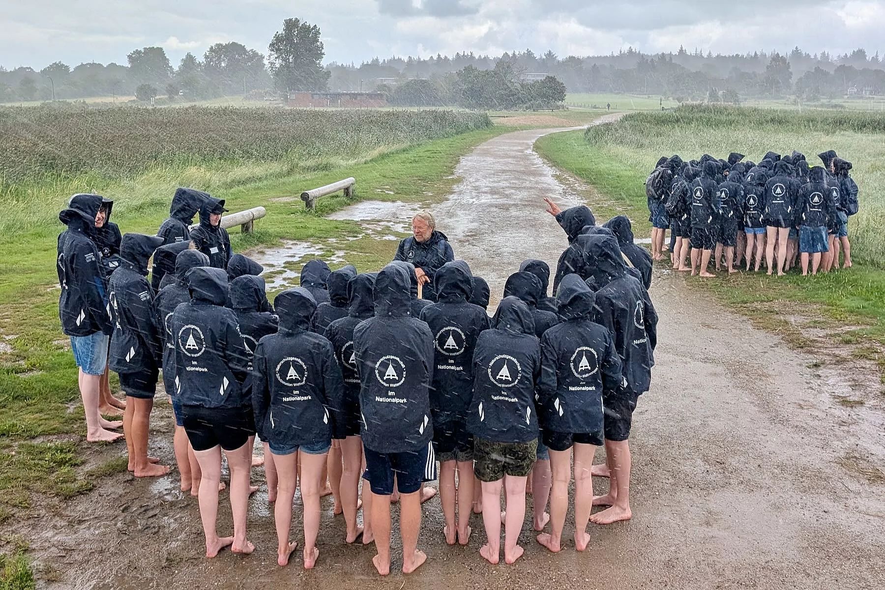 Zwei Gruppen Jugendlicher mit dunkelblauen Regenjacken, stehen bei einem Regenschauer im Freien und lauschen einem Vortrag.