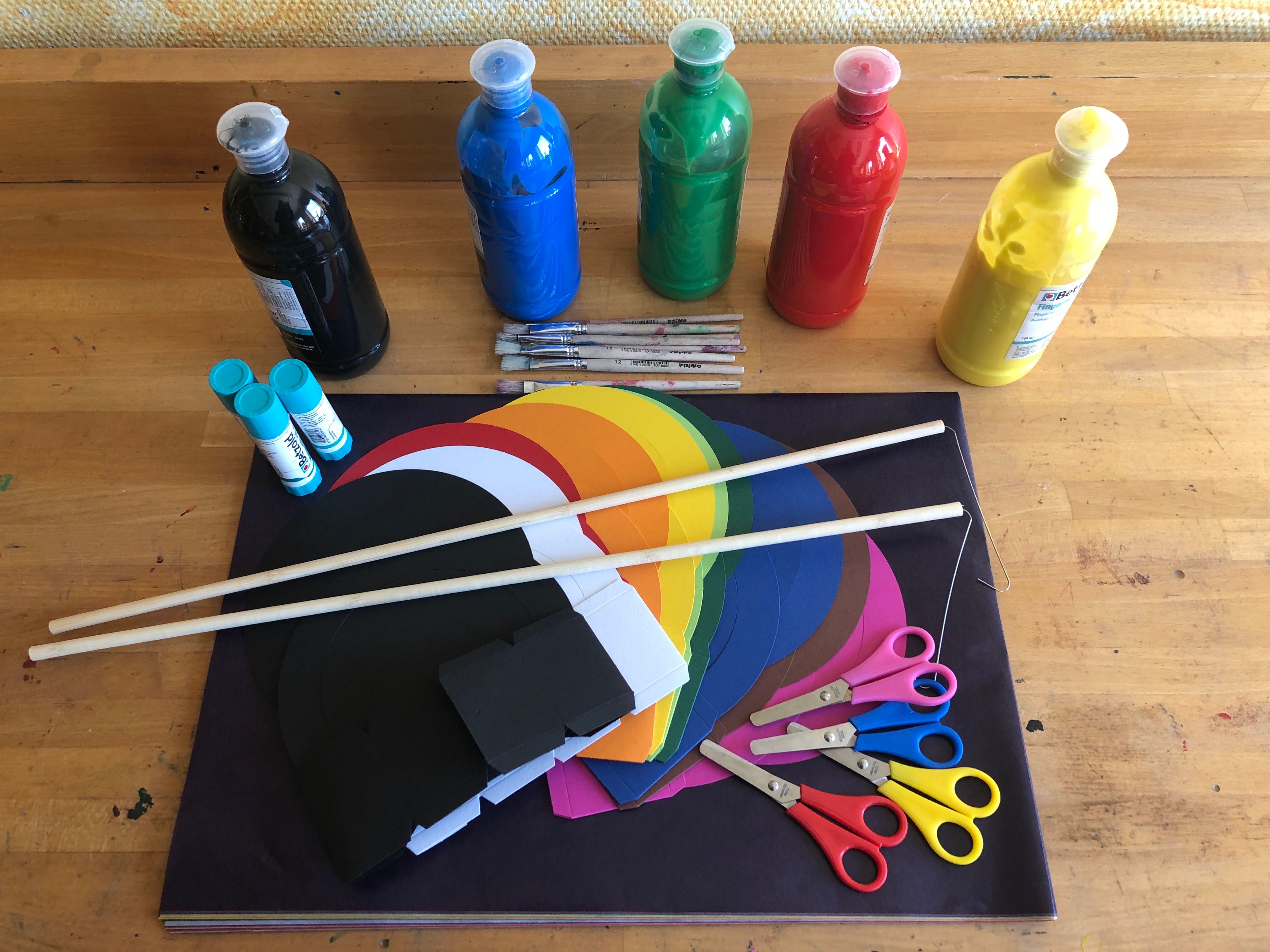 Bastelmaterial für die eigene Laterne, liegt präsentiert auf einem Tisch. Flaschen mit bunter Plakafarbe sowie Pinsel, Klebestifte, buntes Laternenpapier, Papierscheren und Laternenstöcker zum Aufhängen der selbstgebastelten Laterne.
