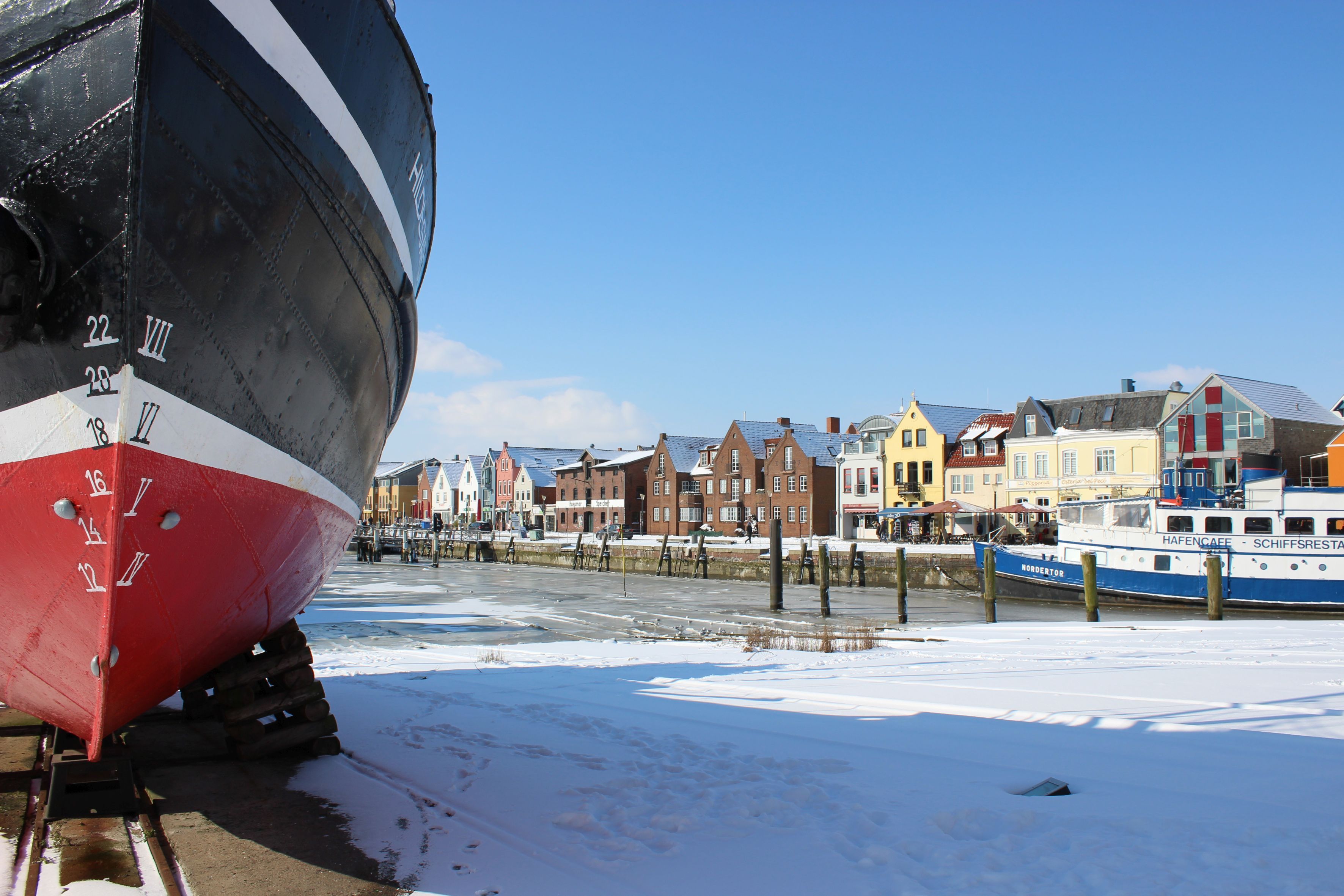 Der Husumer Hafen im Winter. Von der alten Slipanlage der ehemaligen Husumer Werft, auf dessen Gelände heute das neue Rathaus steht, schweift der Blick rechts an dem alten Tonnenleger "Hildegard" vorbei in Richtung alter Husumer Speicher und den Gebäuden auf der Nordseite des Binnenhafens. Rechts ist das Restaurantschiff "Nordertor" zu sehen ein alter Flensburger Ausflugsdampfer. Schnee liegt auf den Landflächen und der Hafen hat eine durch die Tide gebrochene Eisschicht auf dem Wasser.