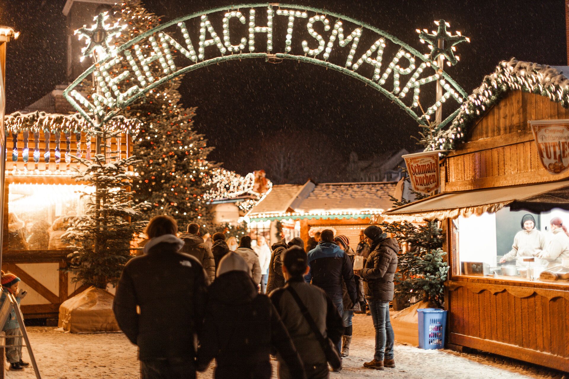 Der westliche Eingang zum Husumer Weihnachtsmarkt mit typischen Mutzen- und Gebrannte Mandelnbuden. Über zwei Buden ragt in einem großzügigen Bogen der Schriftzug "Weihnachtsmarkt". Weiße Glühbirnen bildern dabei die Buchstaben.