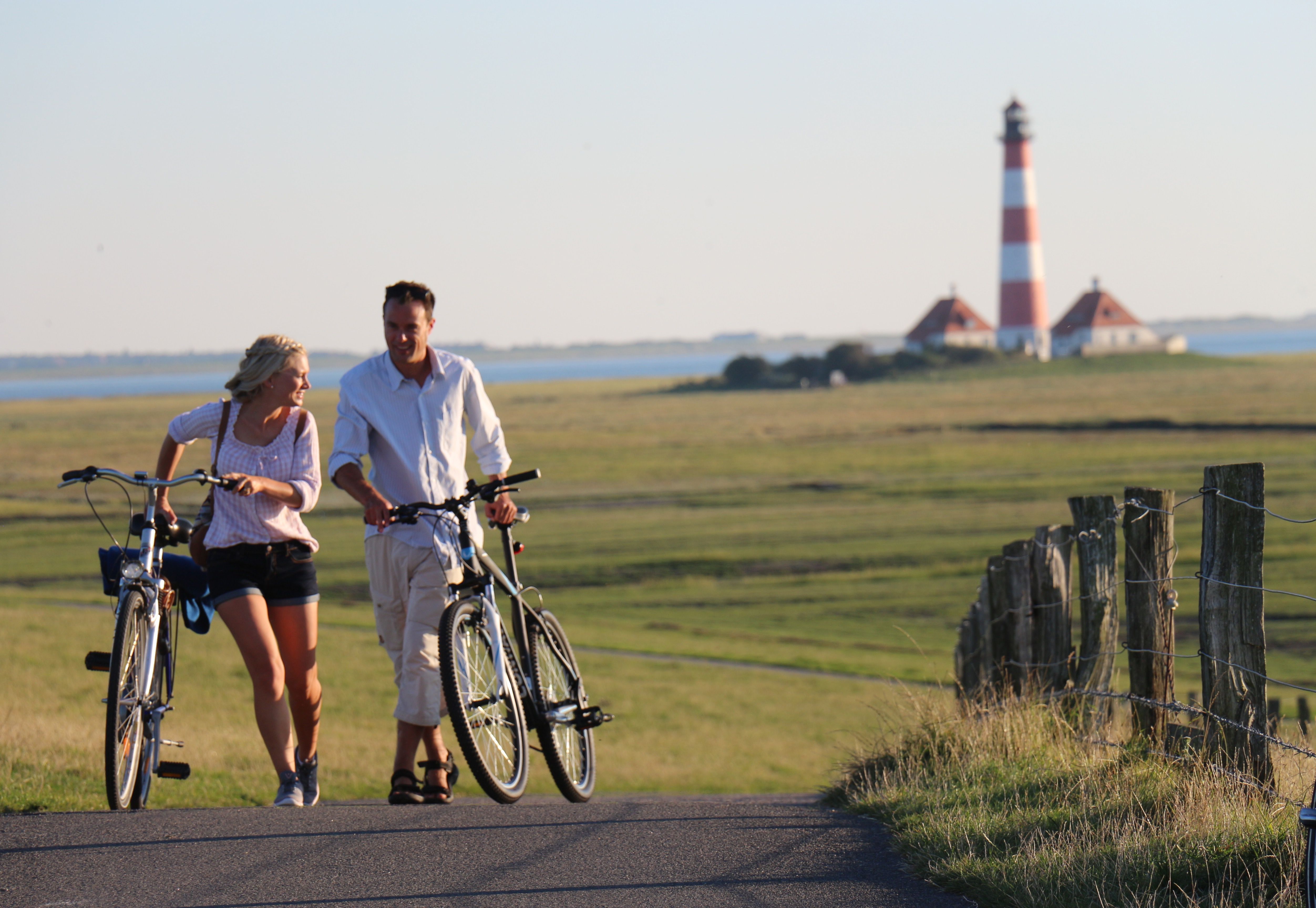 Zwei Radfahrer schieben ihre Fahrräder durch die Salzwiesen, im Hintergrund ist der Westerhever Leuchtturm zu sehen