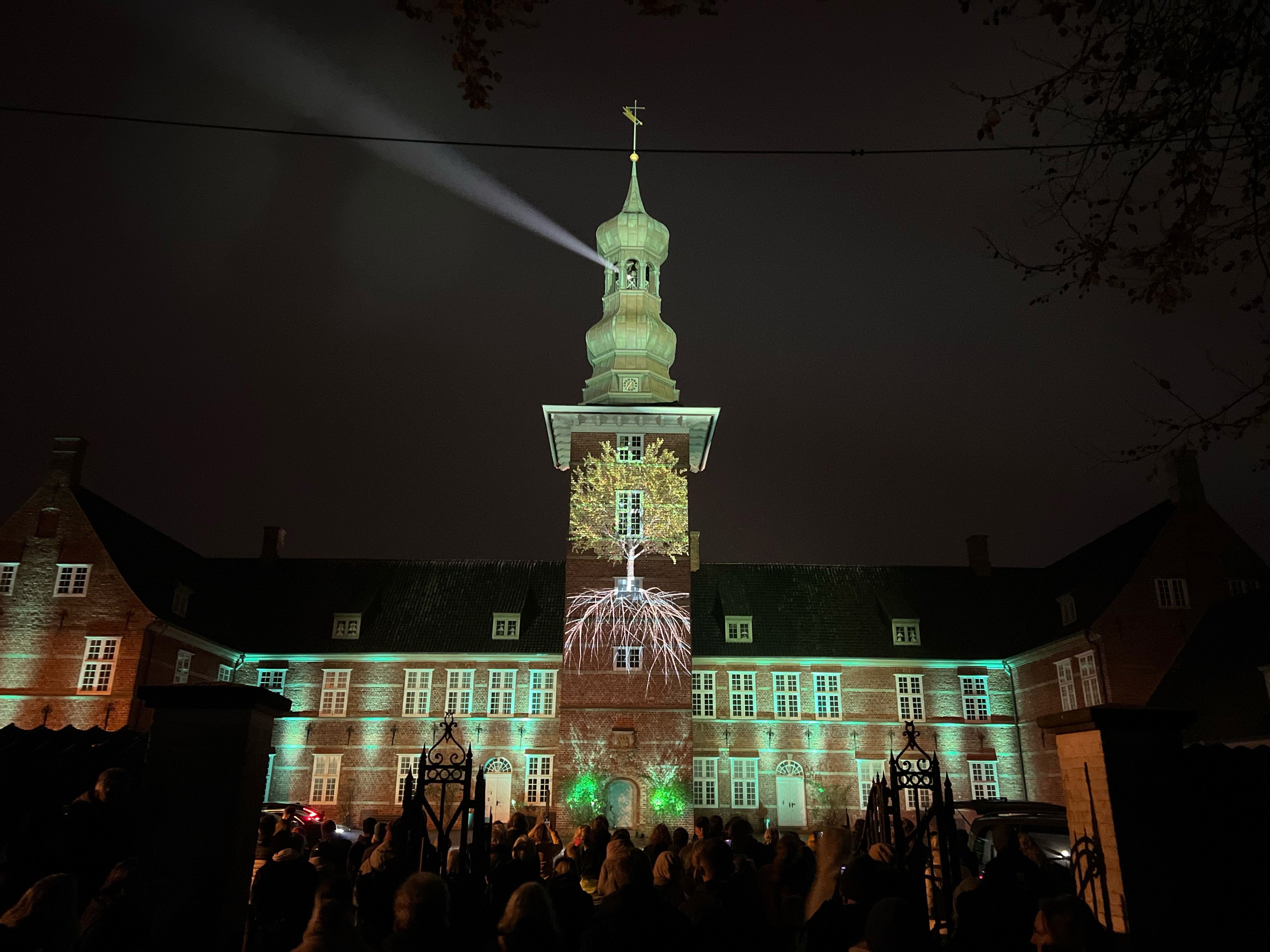 Blick durch das Schlosstor auf das illuminierte "Schloß vor Husum" welches von einer Menschenmenge bestaunt wird.. Auf der Schlossturmspitze strahlt ein Lichtkegel wie bei einem Leuchtturm in die Nacht.