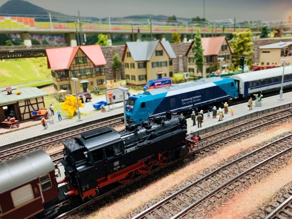 Verschiedene Modellzüge stehen an einem kleinen Bahnhof, an dem Passagiere warten. Im Hintergrund stehen kleine Häuser