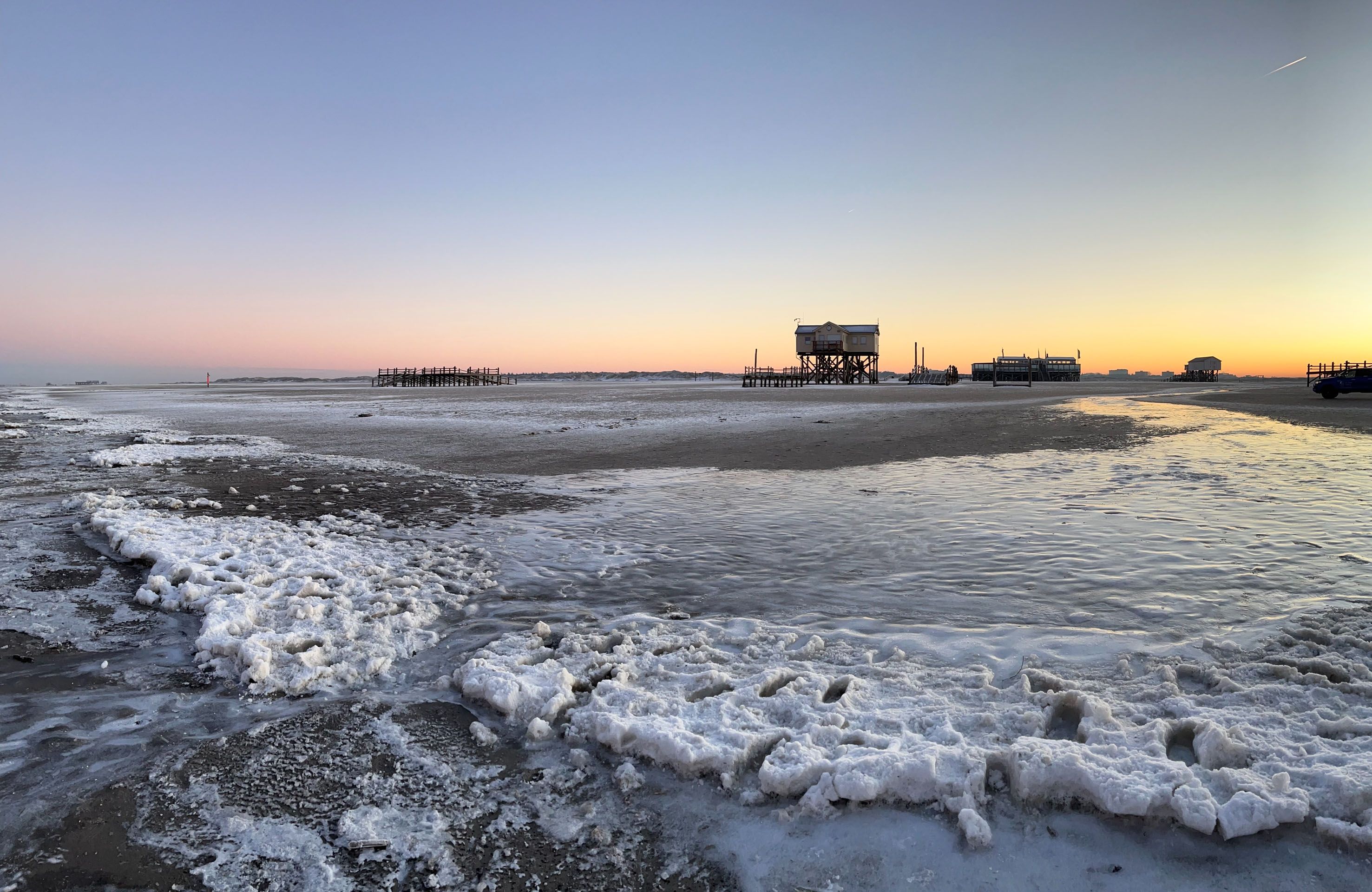 Der Blick geht über den teilweise vereisten Sandstrand von St. Peter-Ording in Richtung Osten. Zusehen sind ein paar der berühmten Pfahlbauten und Pfahlterassen sowie der gerade stattfindende Sonnenaufgang.