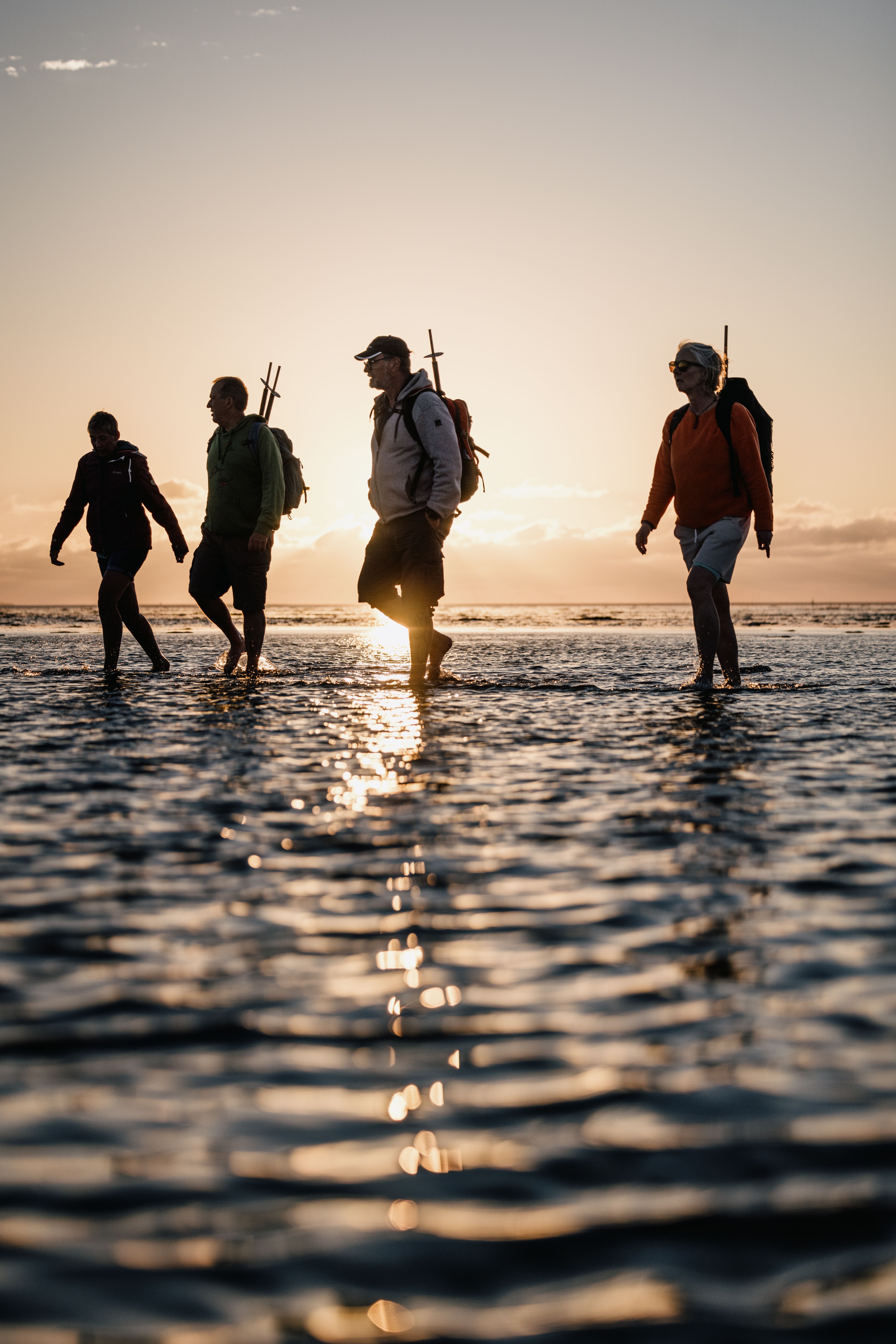 Vier Personen laufen mit Ausrüstung durch seichtes Wasser, im Hintergrund geht die Sonne unter