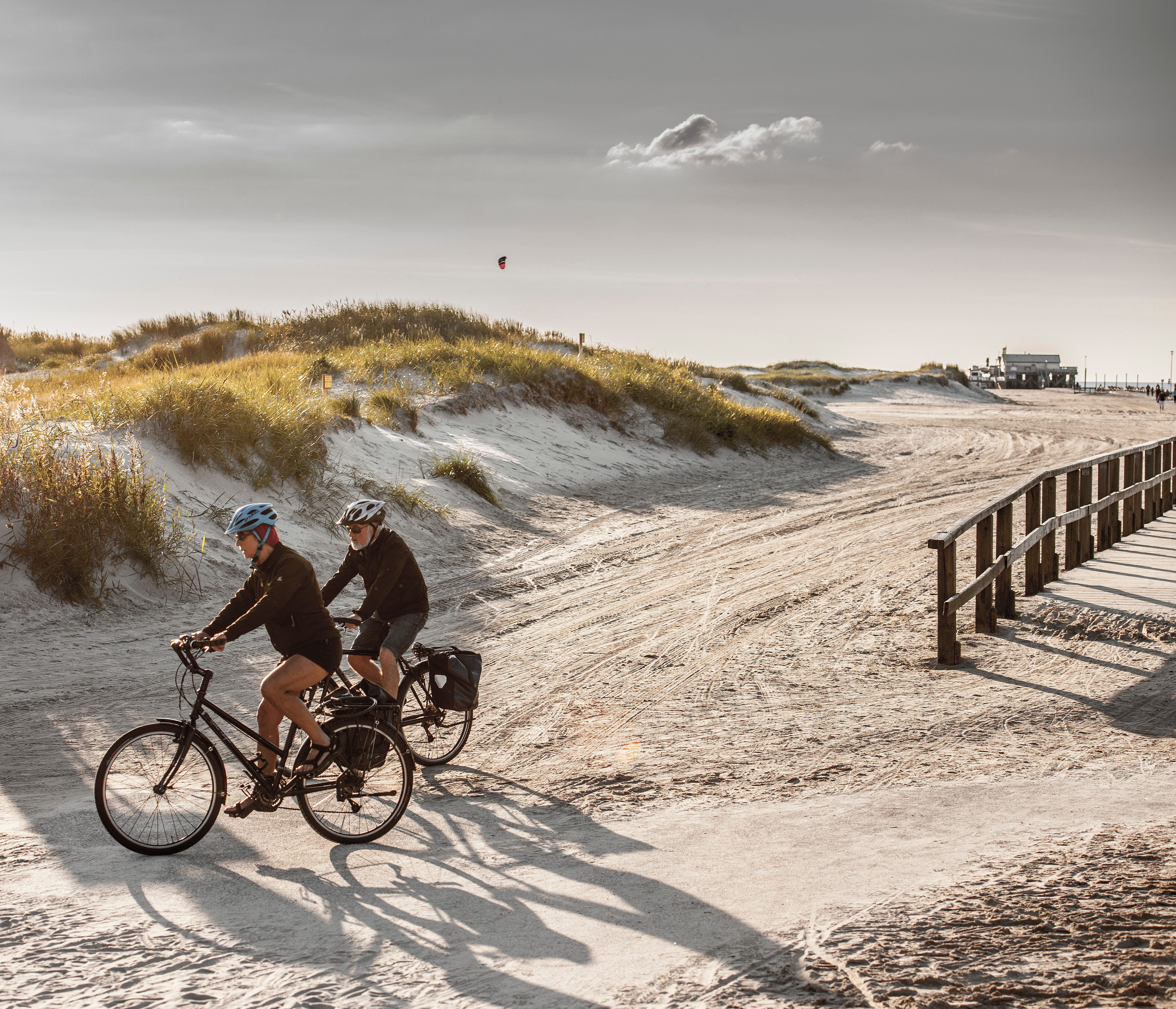 Zwei Radfahrer fahren am Strand von St. Peter-Ording entlang, im Hintergrund sind Dünen und eine Pfahlbaute zu sehen