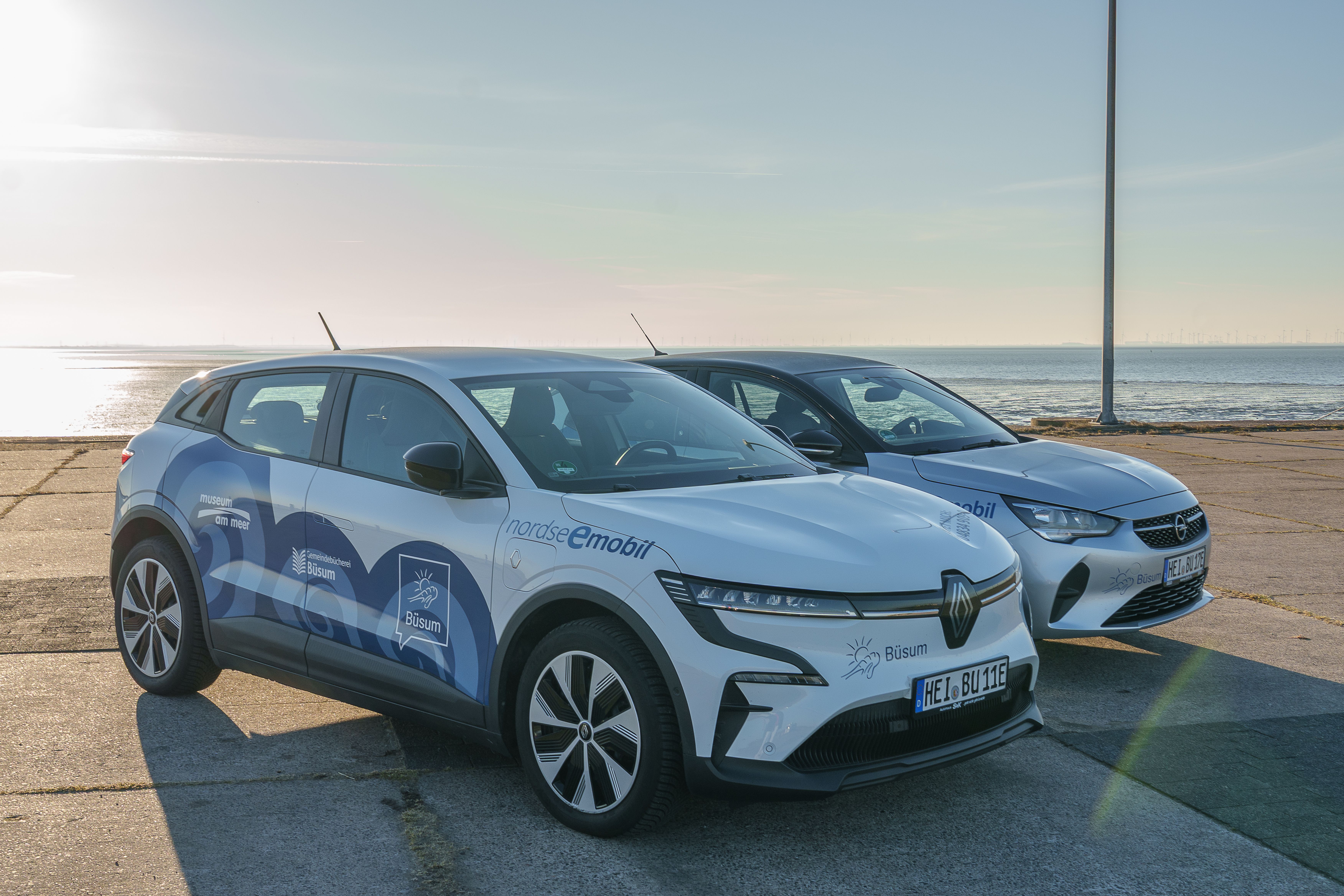 Zwei weiße Nordseemobile (Elektroautos mit blauen Wellendesign) stehen auf der Promenade von Büsum direkt an der Nordsee.