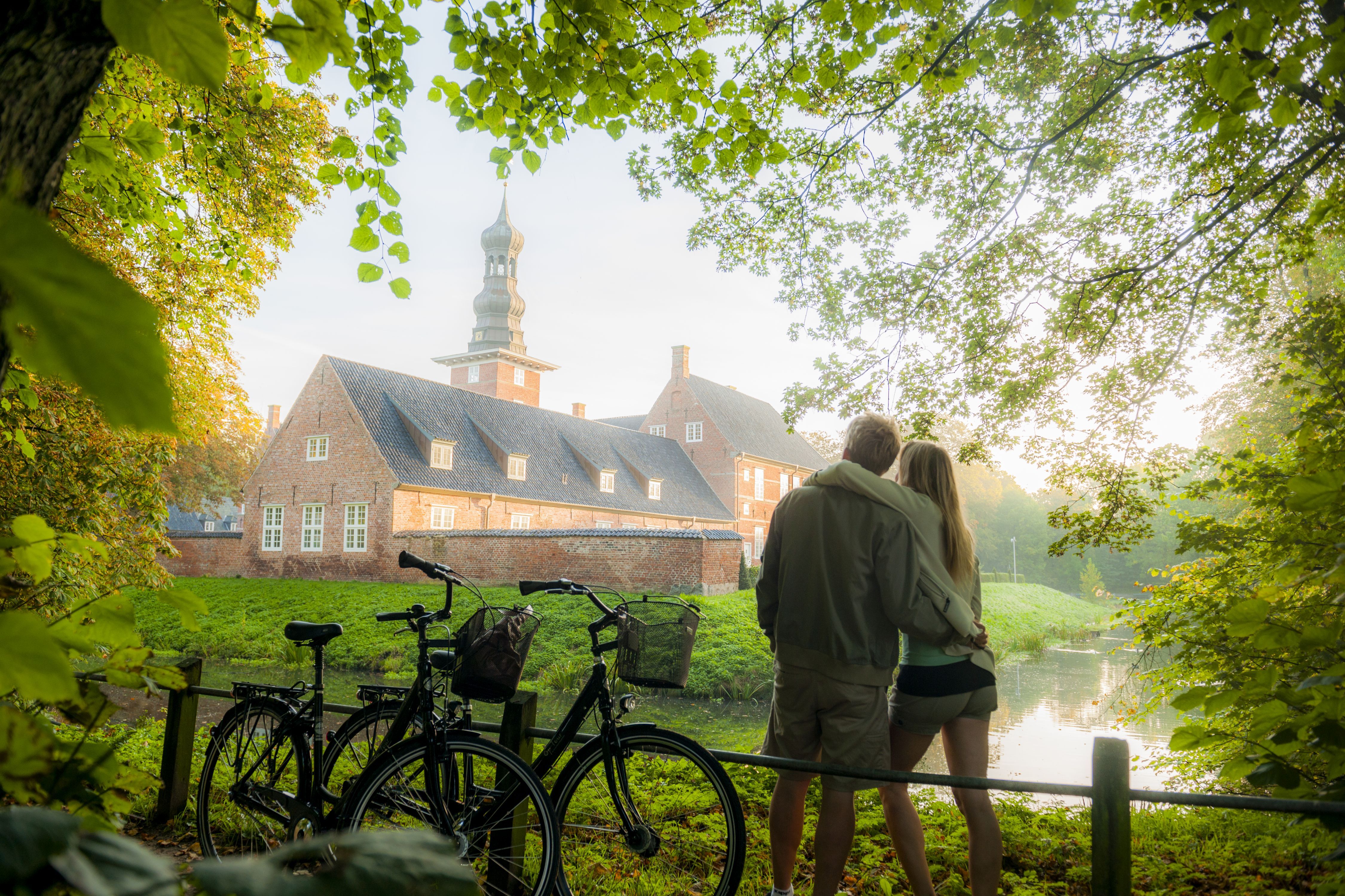 Ein Pärchen steht an einem sonnigen Tag mit ihren Fahrrädern vor dem Schloss in Husum. Sie blicken über den Wassergraben zum Schloss hinüber.