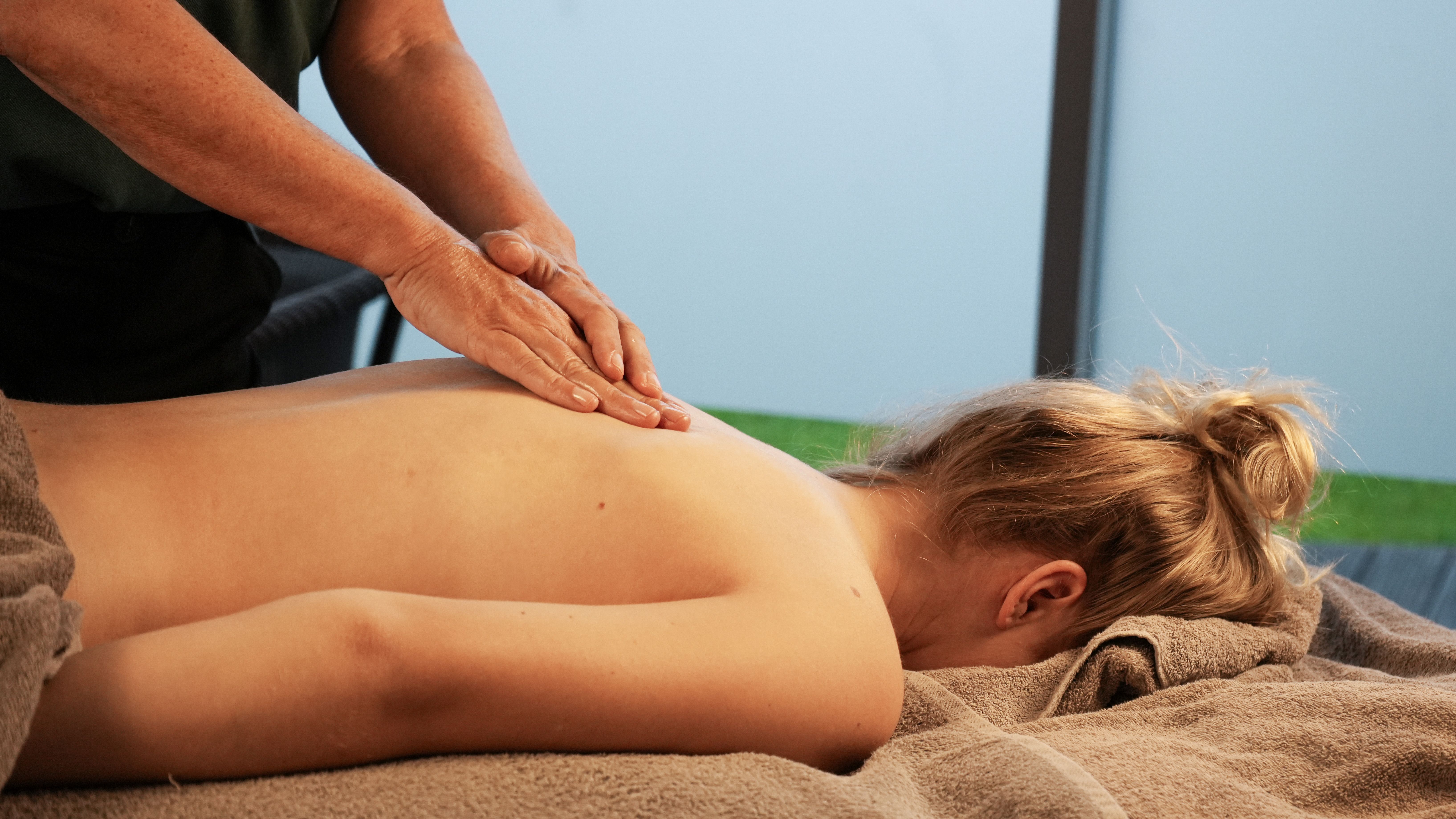 Eine Frau mit blonden langen Haaren, liegt mit dem Gesicht nach unten, unbekleidet auf einer Wellnes-Behandlungsliege, während sie von einem Masseur zwischen den Schulterblättern massiert wird.