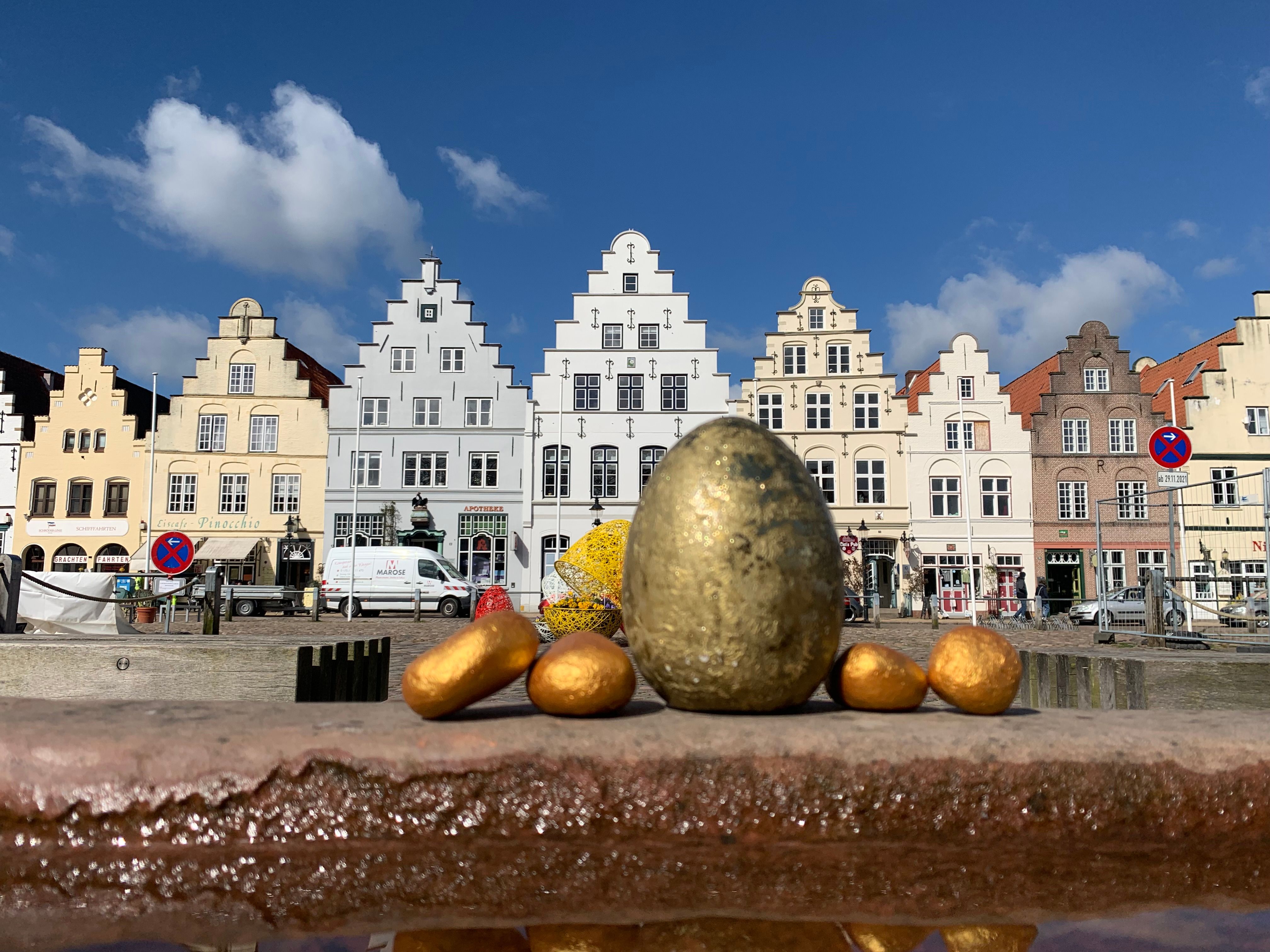 Ein großes goldenes Ei, steht auf eine Steinkante vor der Shilouette des Friedrichstädter Marktplatzes.