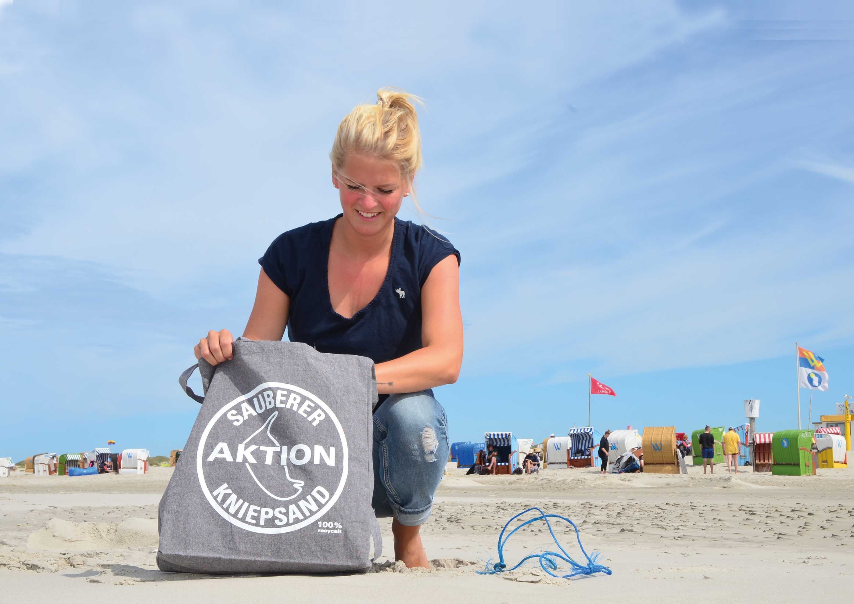 Eine Frau kniet im Amrumer Kniepsand, vor sich eine graue Tasche mit der Aufschrift "Aktion sauberer Kniepsand"