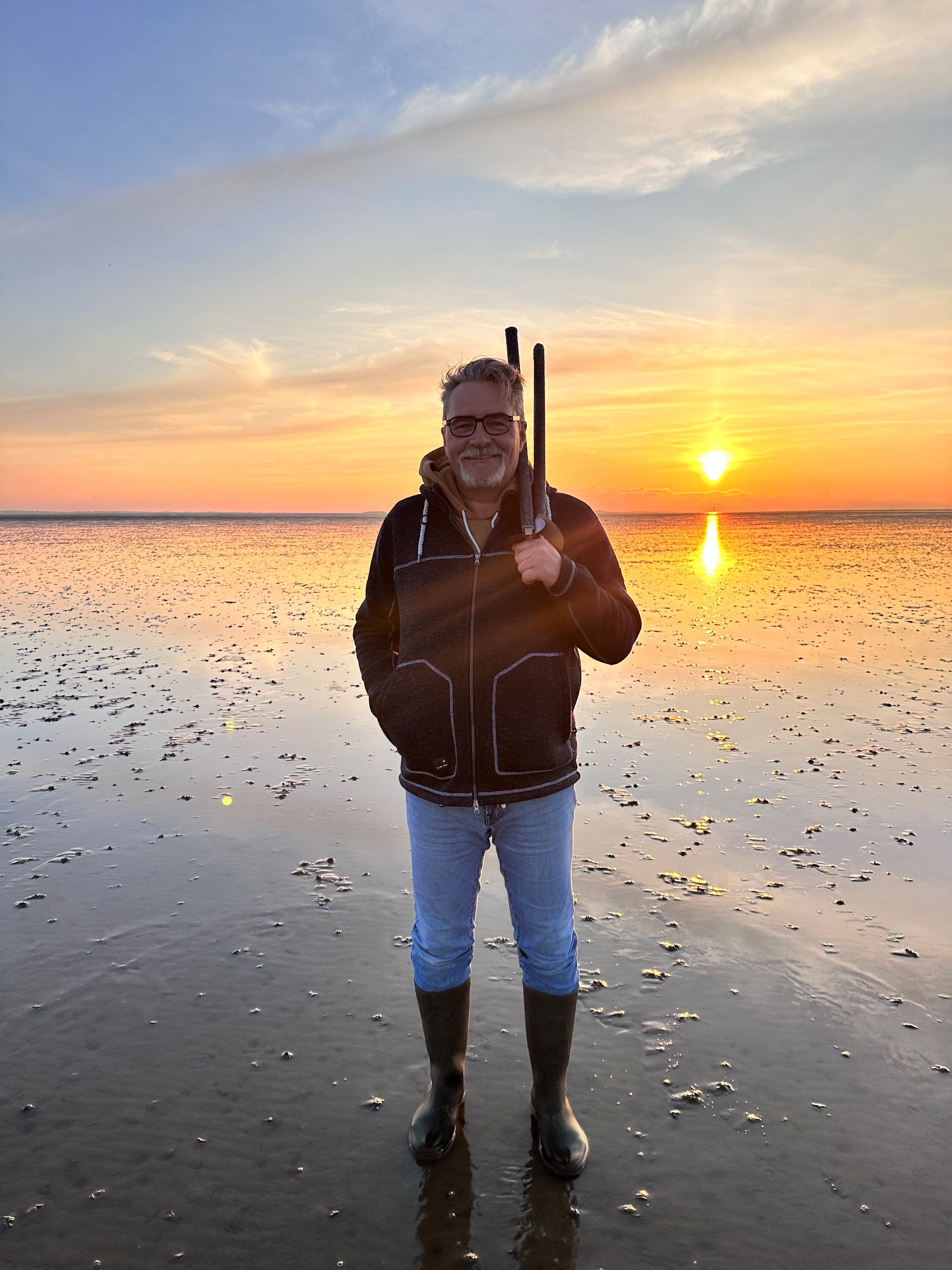 Ein Wattführer steht bei Sonnenuntergang mit geschulterter Mistgabel im Watt vor Dagebüll.