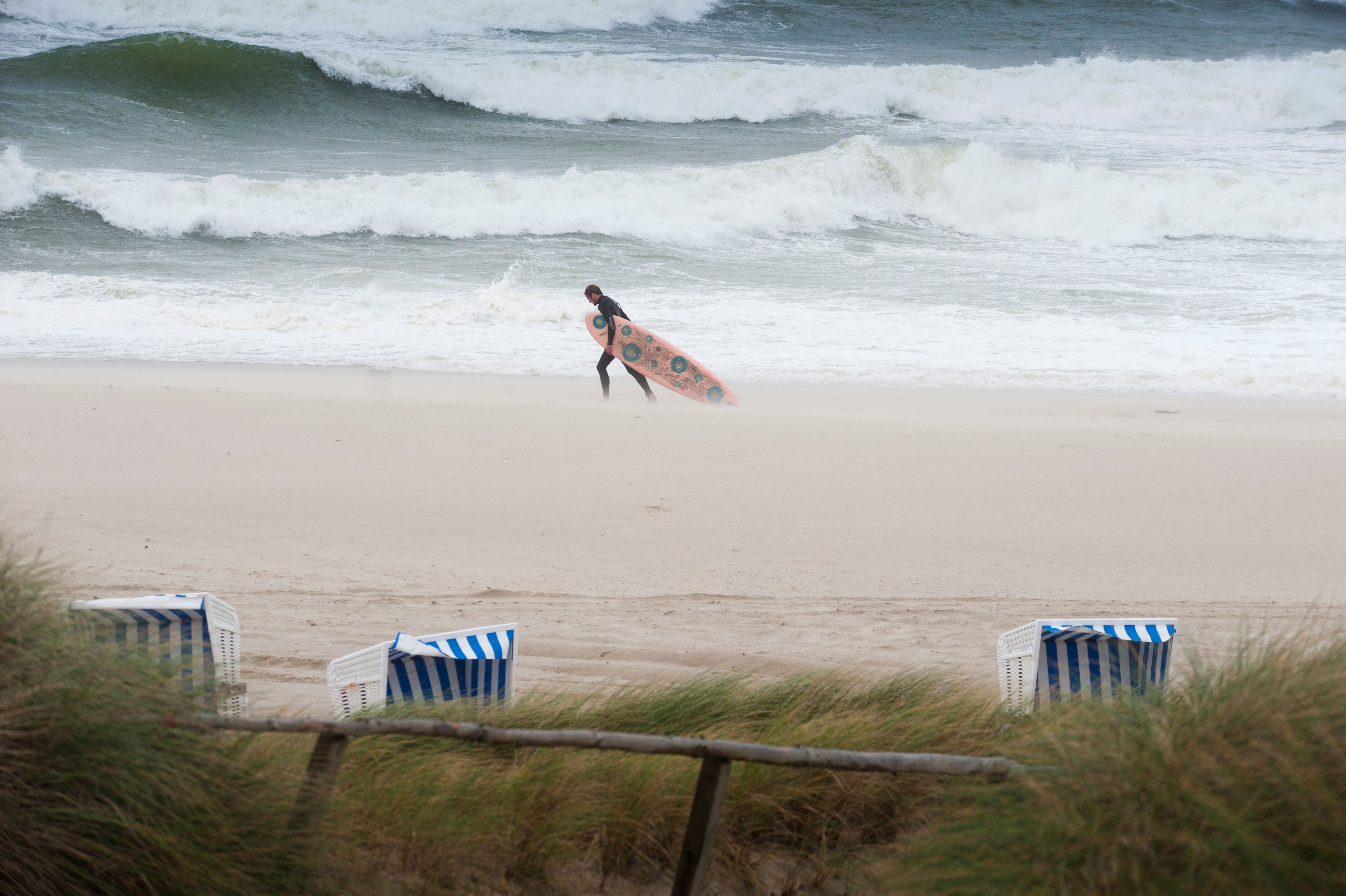 Stürmische See, starker wind von links nach rechts. Der Blick geht durch Dünengras vorbei an Dächern von Strandkörben auf den Strand, wo ein Wellenreits im schwarzen Neoprenanzu, sein Surfboard gegen den Wind schleppt, dabei ist das Brett mit dem vorderen Teil unter seinem Arm getragen und der untere Teil schleift auf dem Sandstrand. Hinter dem Surfer brechen sich die Wellen.